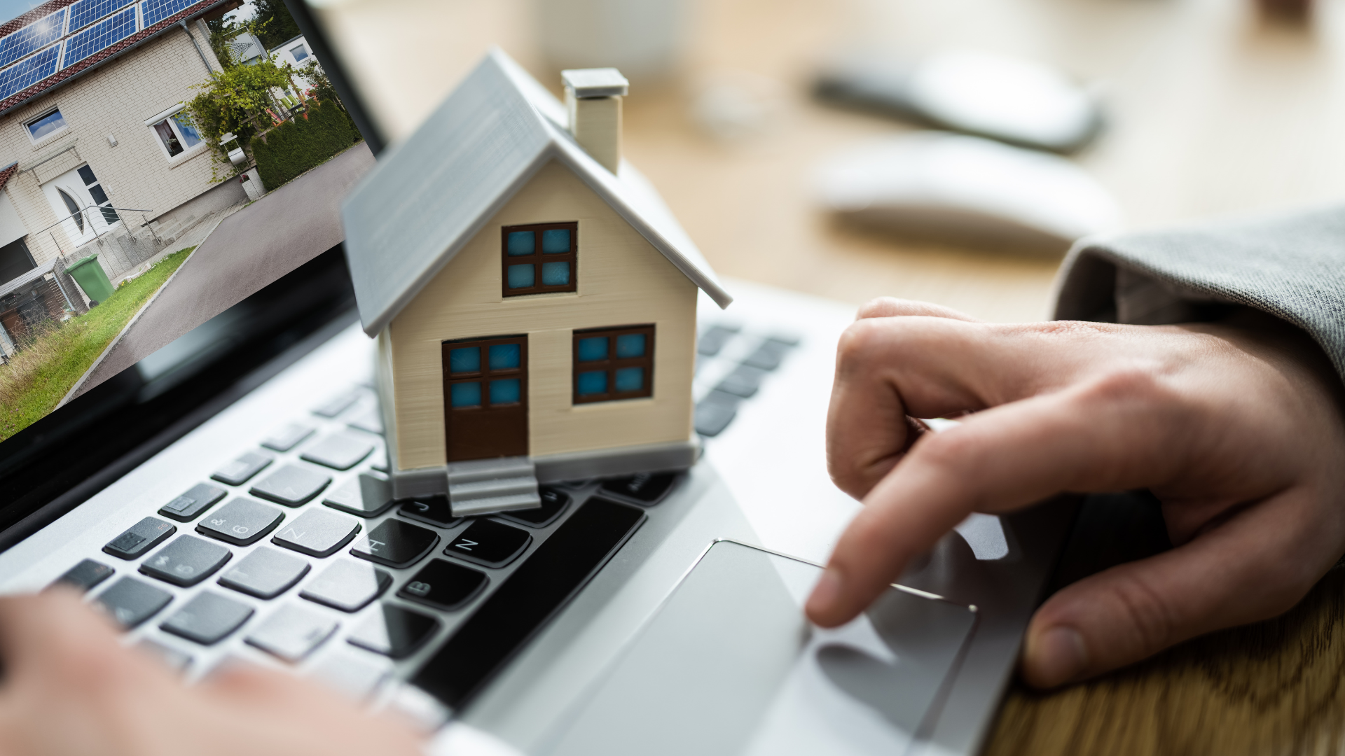 A person using a laptop with a small model house placed on the keyboard, and a real house on a computer screen in the background.