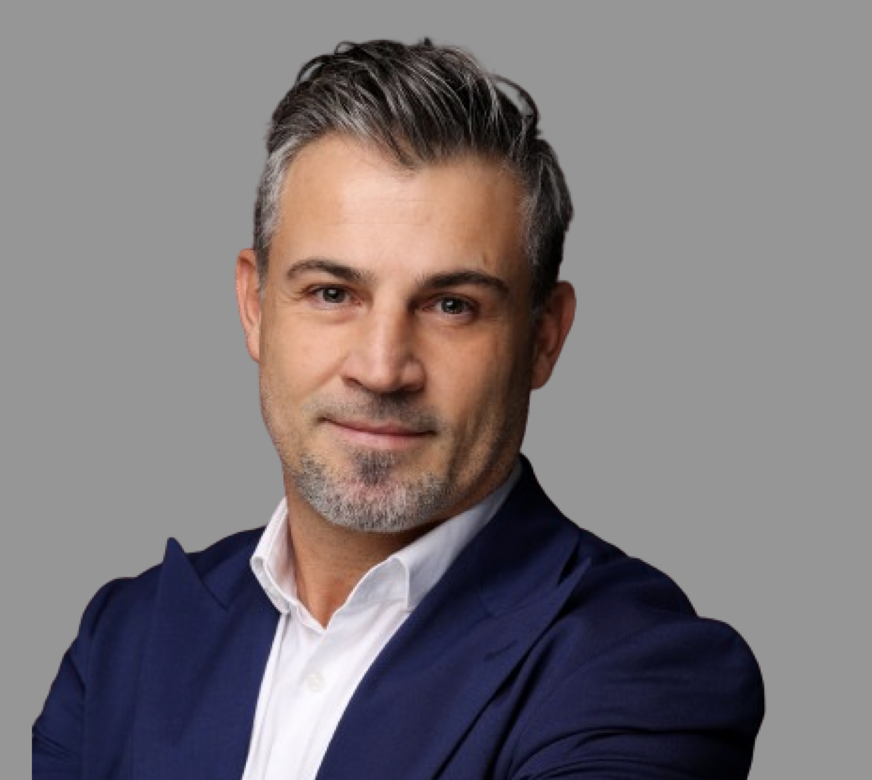 Headshot of a middle-aged man with gray hair and beard, wearing a dark suit and white shirt, smiling slightly against a gray background.