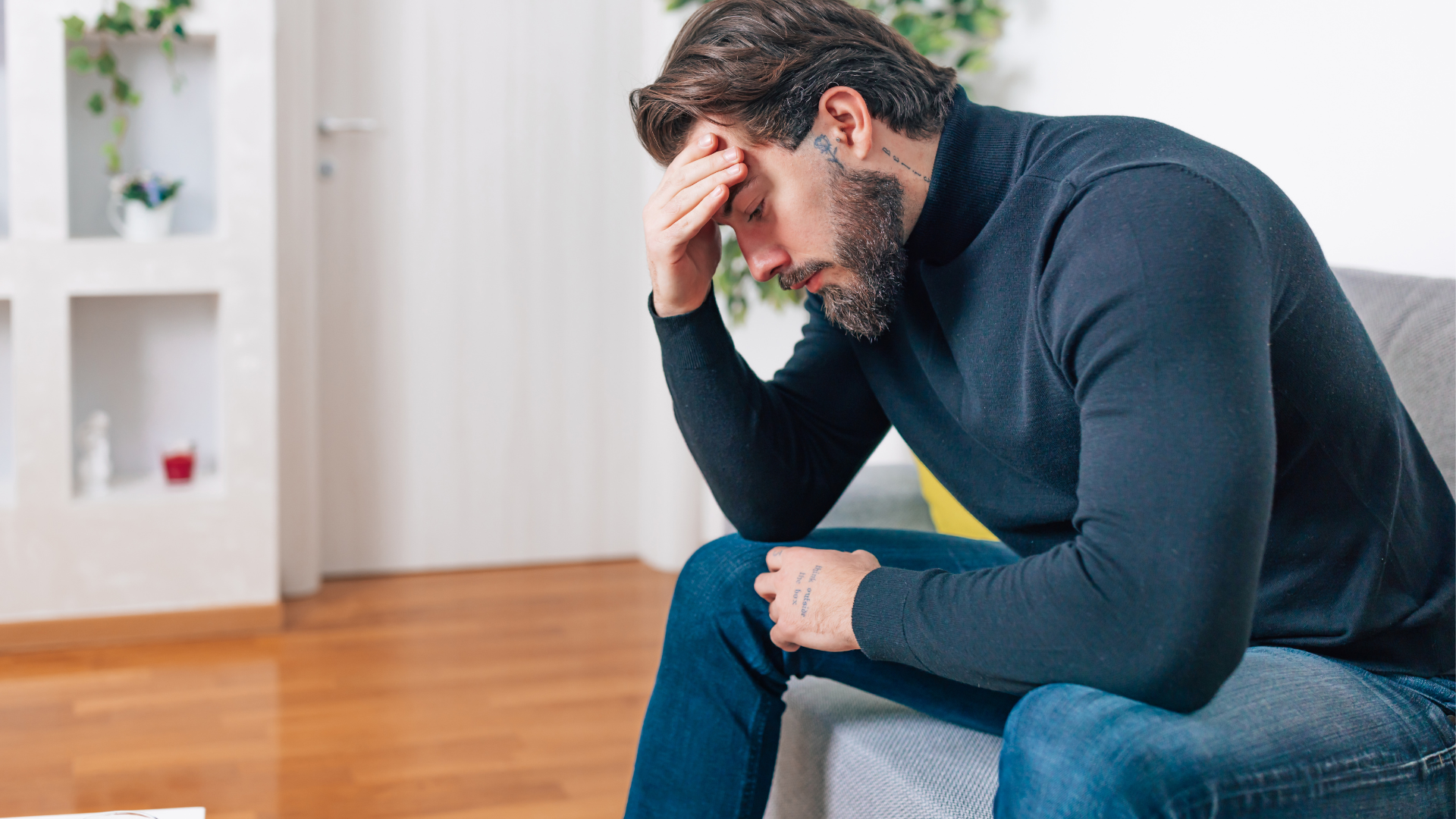 A man sitting on a couch holding his head with his right hand, looking down with a pained expression, in a room with wooden floors and decorative shelves in the background.