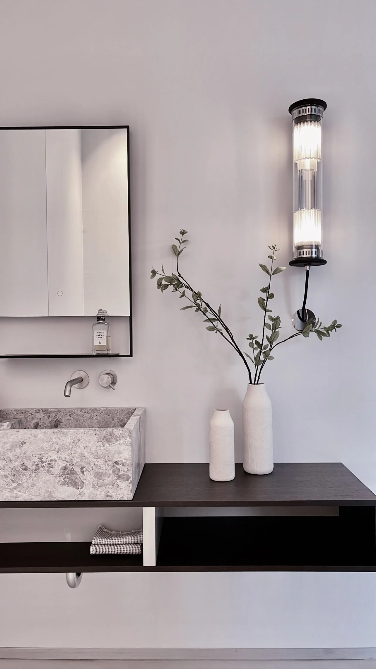 Une pièce de salle de bain minimaliste avec un lavabo en pierre, un miroir, des vases blancs avec des branches d'olivier, et un éclairage mural