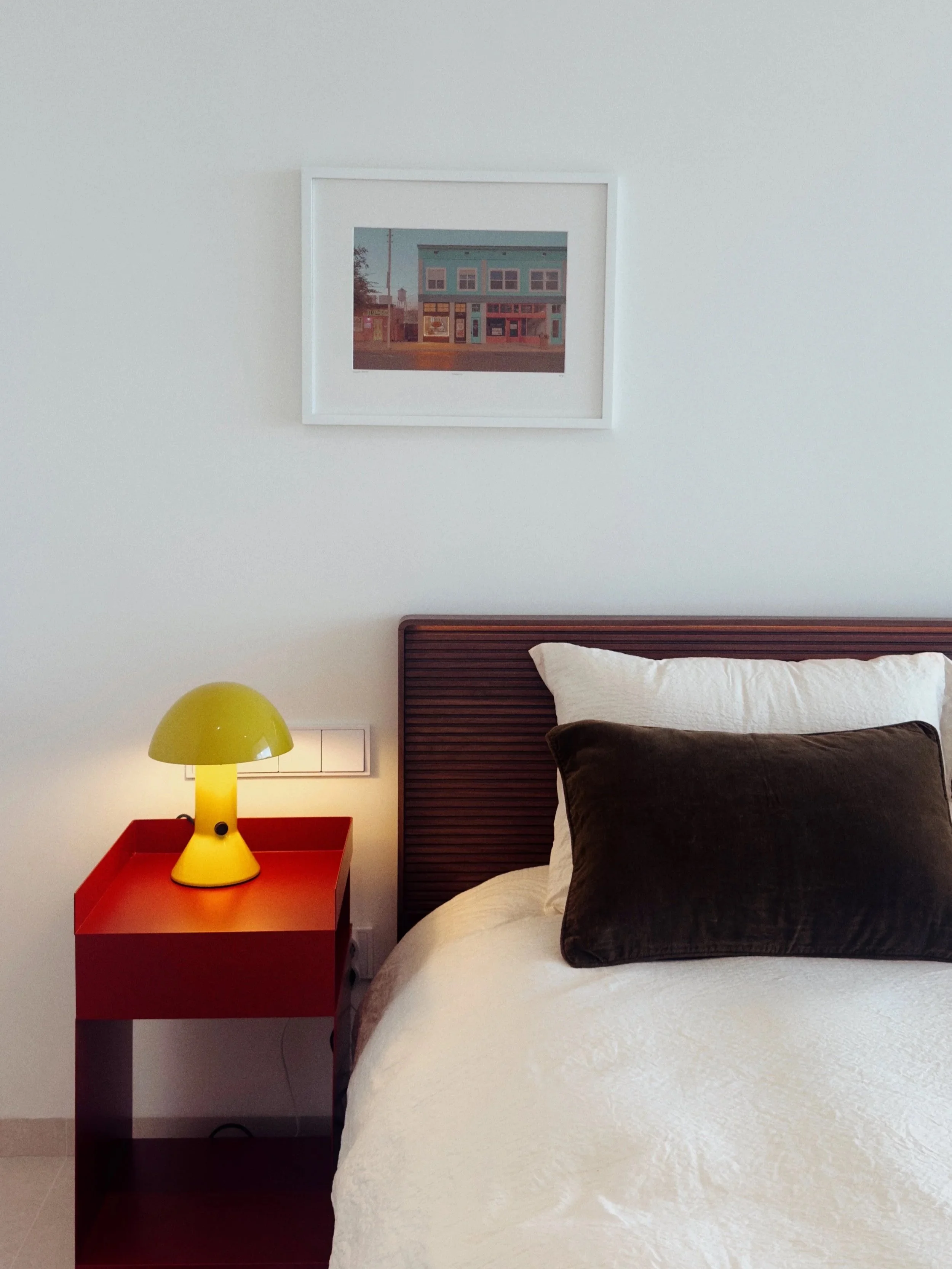 Chambre avec lit blanc, oreiller noir, table de chevet rouge, lampe verte, tableau sur le mur, et prise électrique.
