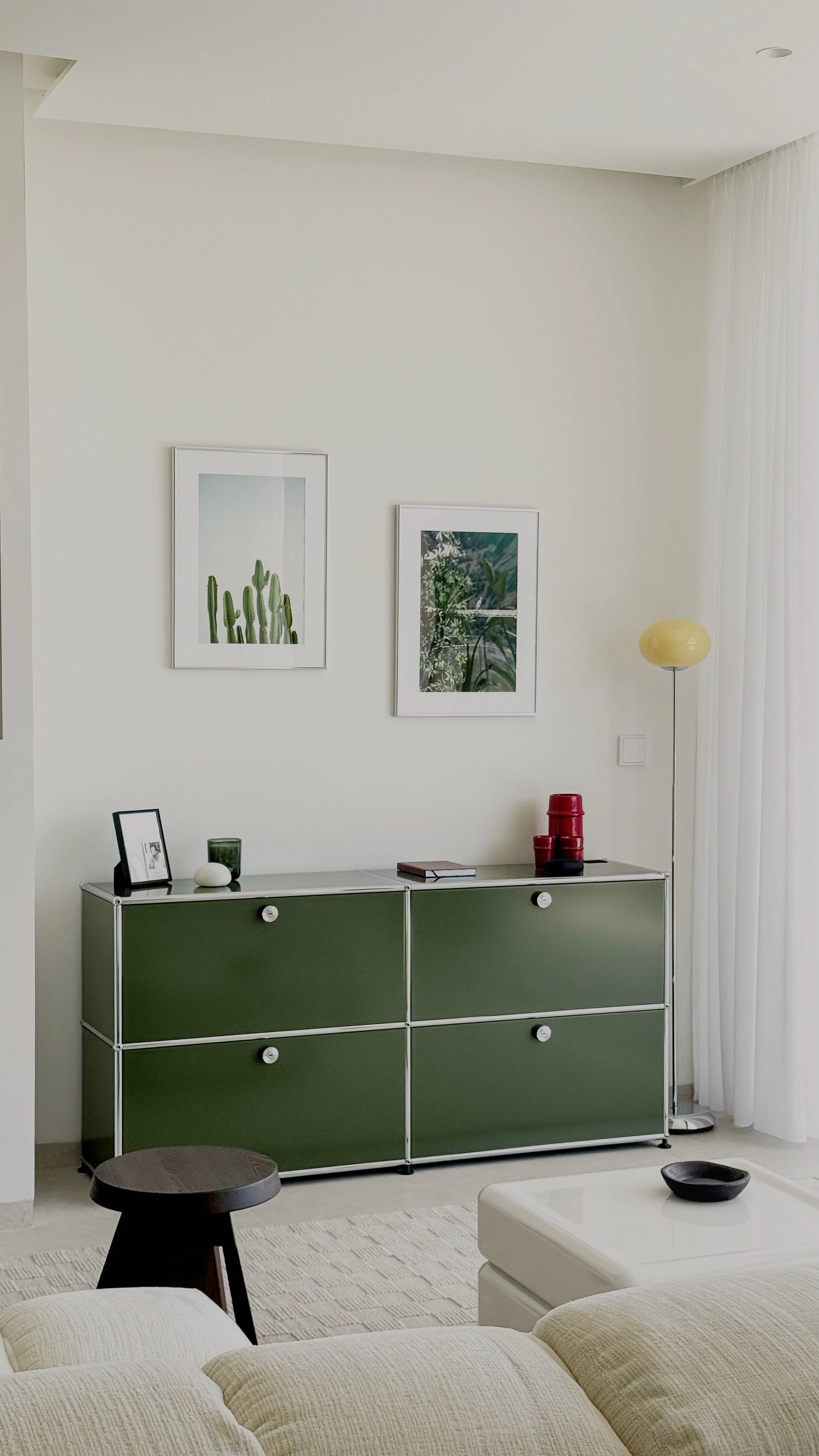 Intérieur moderne avec un meuble vert, deux tableaux avec des images de plantes, une lampe sur pied, et une décoration minimaliste.