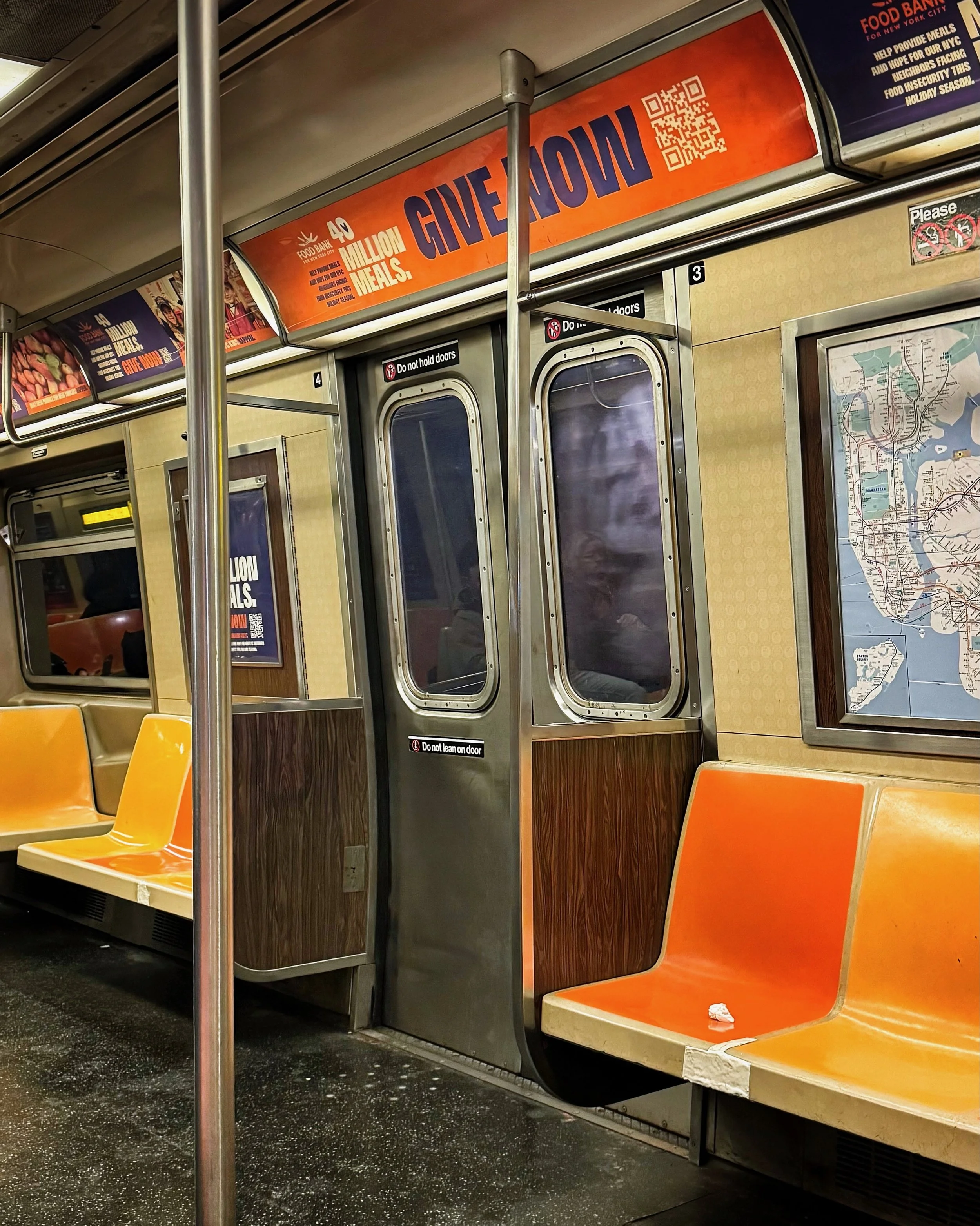 Intérieur d’un métro newyorkais avec des sièges oranges ou jaunes, une carte du métro sur le mur, et des affichages publicitaires en anglais. La porte du métro est fermée.