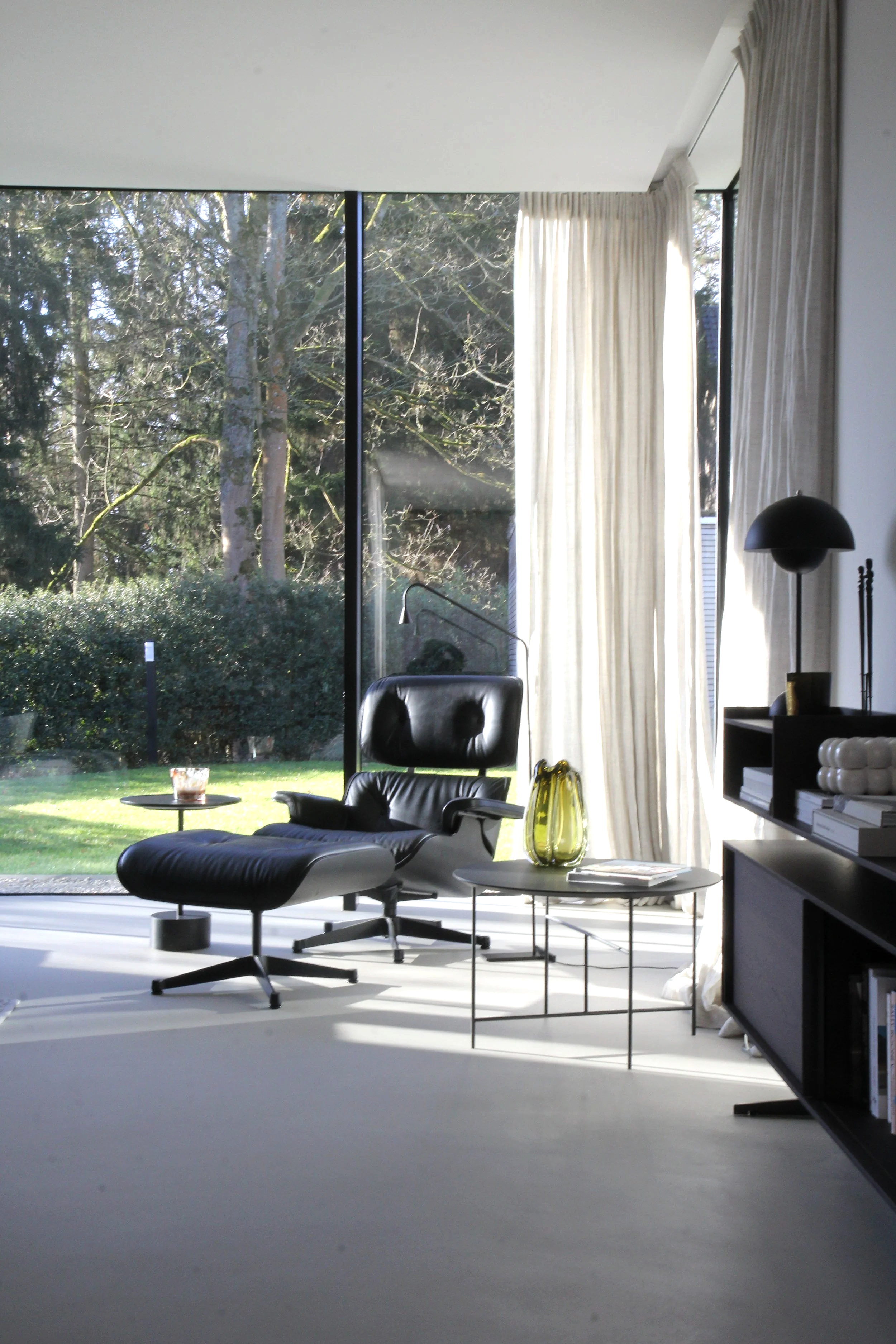 Salon moderne avec un fauteuil Eames Lounge Chair en ottoman en cuir noir, une table basse avec une vase verte, grande baie vitrée avec rideaux gris clairs en lin, vue sur un jardin avec des arbres.