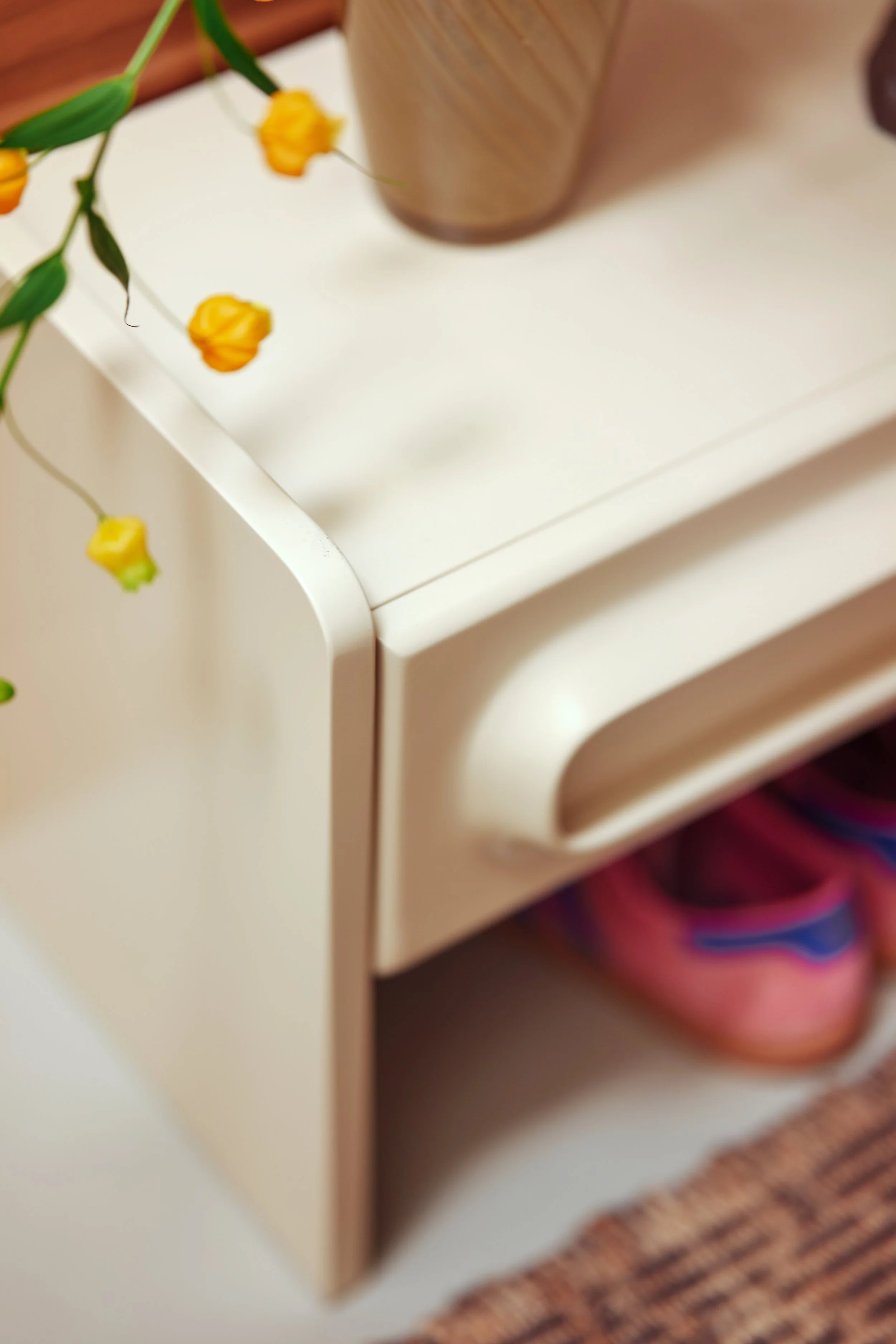 Une petite commode blanche avec une porte entrouverte, une boîte rose à l'intérieur, et une plante verte avec des fleurs jaunes posées dessus.