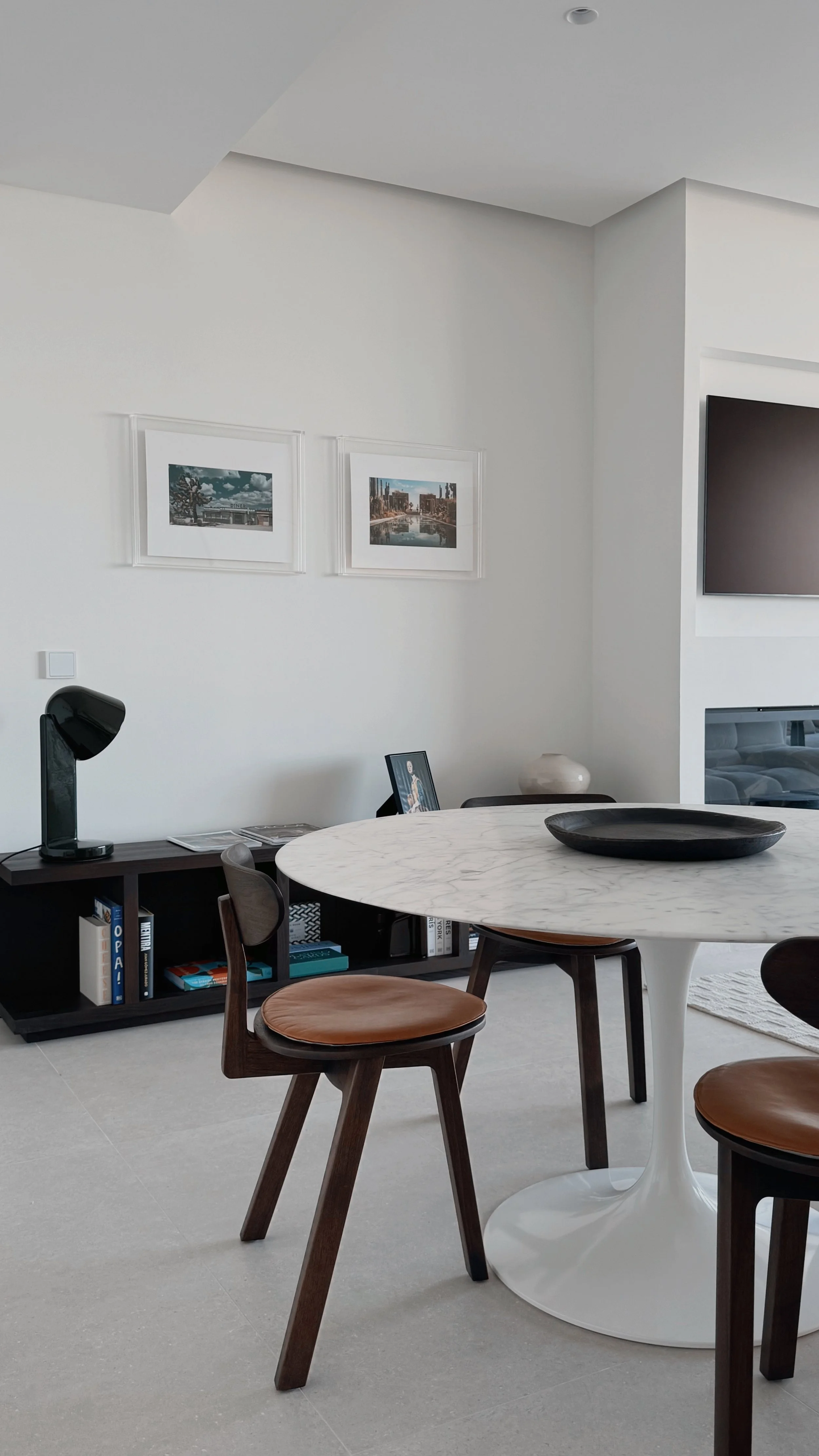 Salle à manger moderne avec table ronde en marbre blanc, chaises en bois avec assises en cuir marron, affiches encadrées sur le mur blanc, meuble noir avec livres et un lampe noire