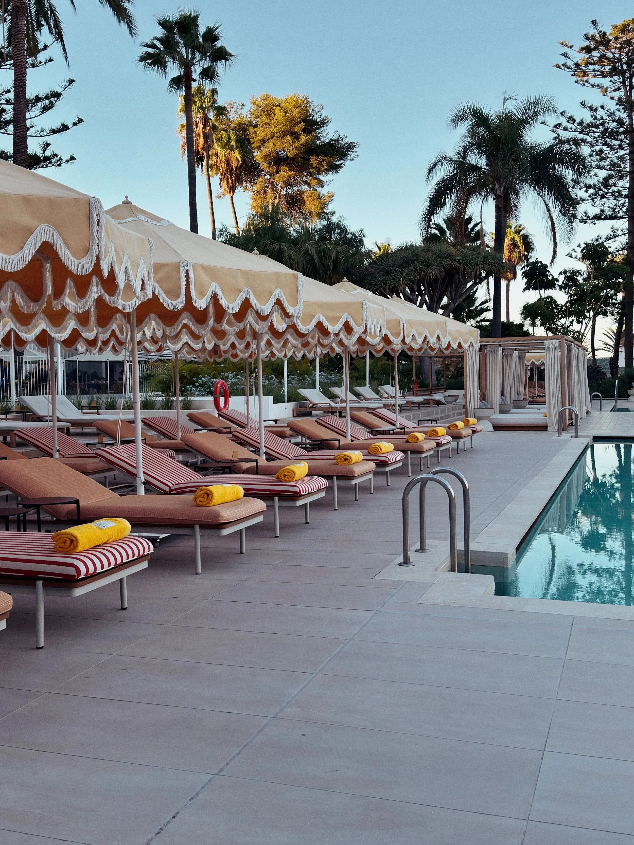 Chaises longues avec des serviettes jaunes pliées, parasols beige avec franges, piscine entourée de palmiers et d'arbres dans un cadre de piscine en plein air