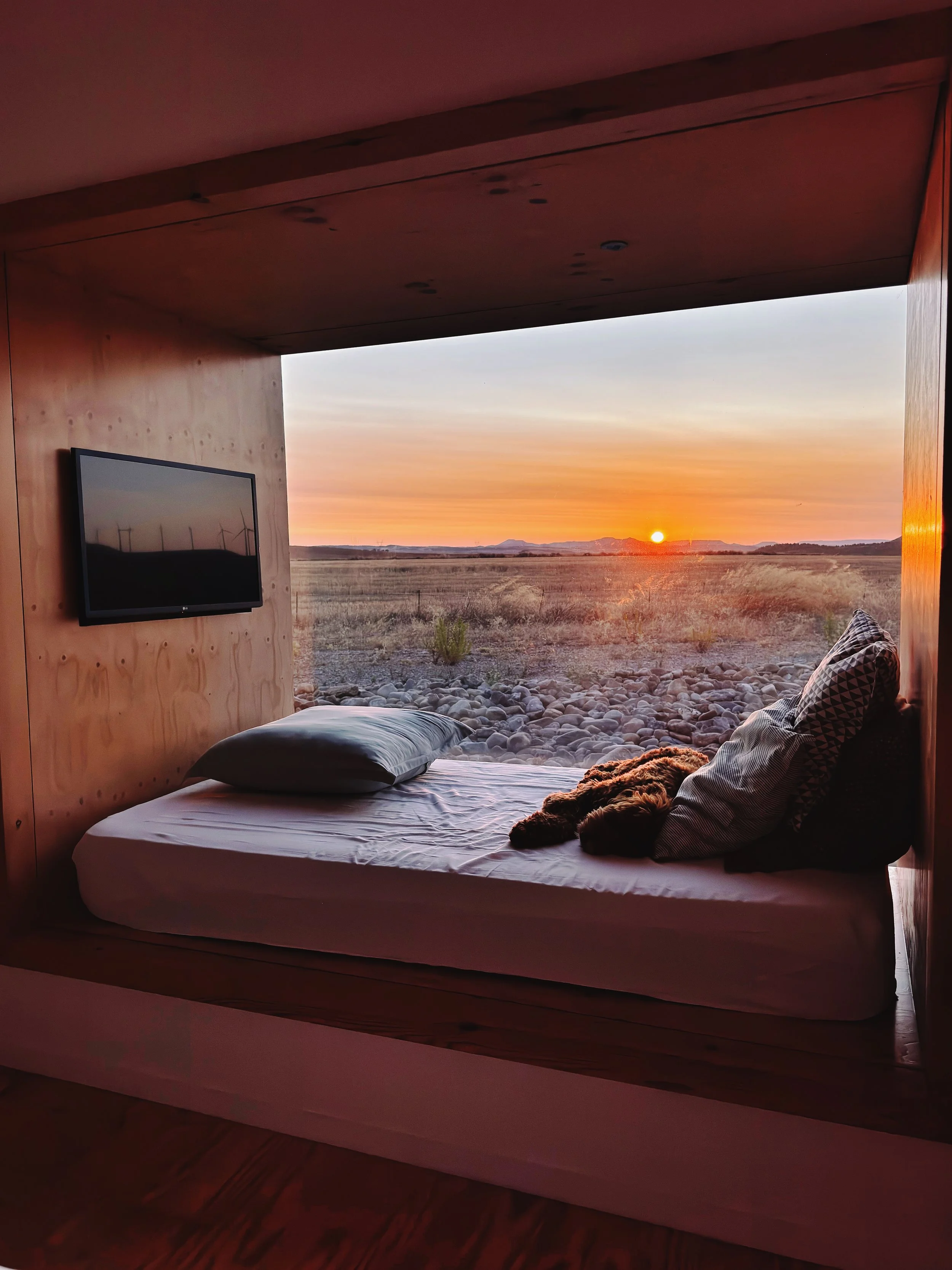 Vue intérieure d'une chambre avec un lit, un canapé, un mur en bois et une grande fenêtre ouverte sur un paysage de champs au coucher de soleil.