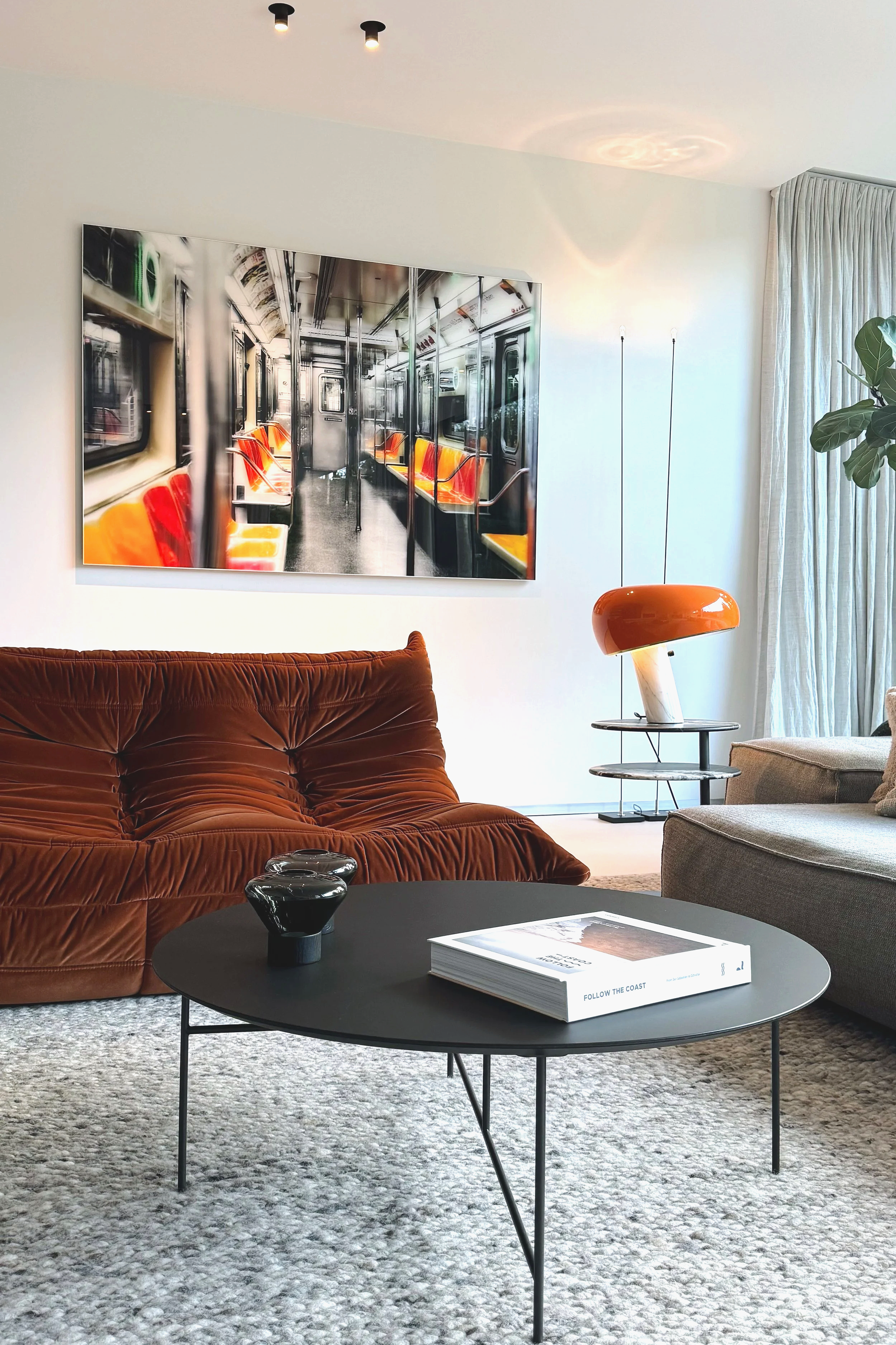 Salon moderne avec un canapé Togo, une table basse noire avec un livre et deux vases noirs, un lampadaire Snoopy, une grande peinture représentant un métro avec des sièges orange et jaunes, et une plante verte près d'une baie vitrée 