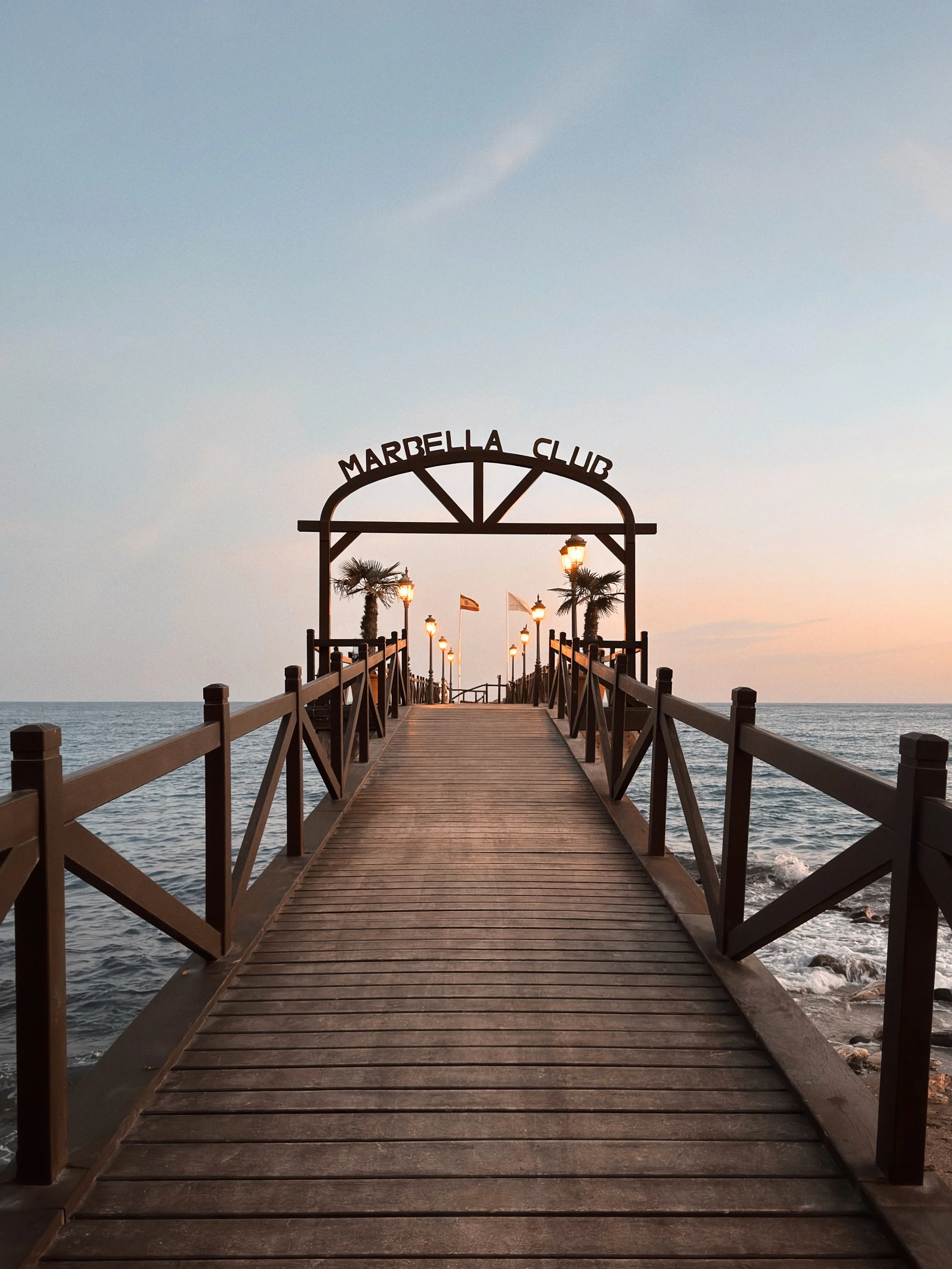 Un pont en bois menant vers la mer, avec des lampadaires allumés, des palmiers et un ciel au coucher du soleil à Marbella.
