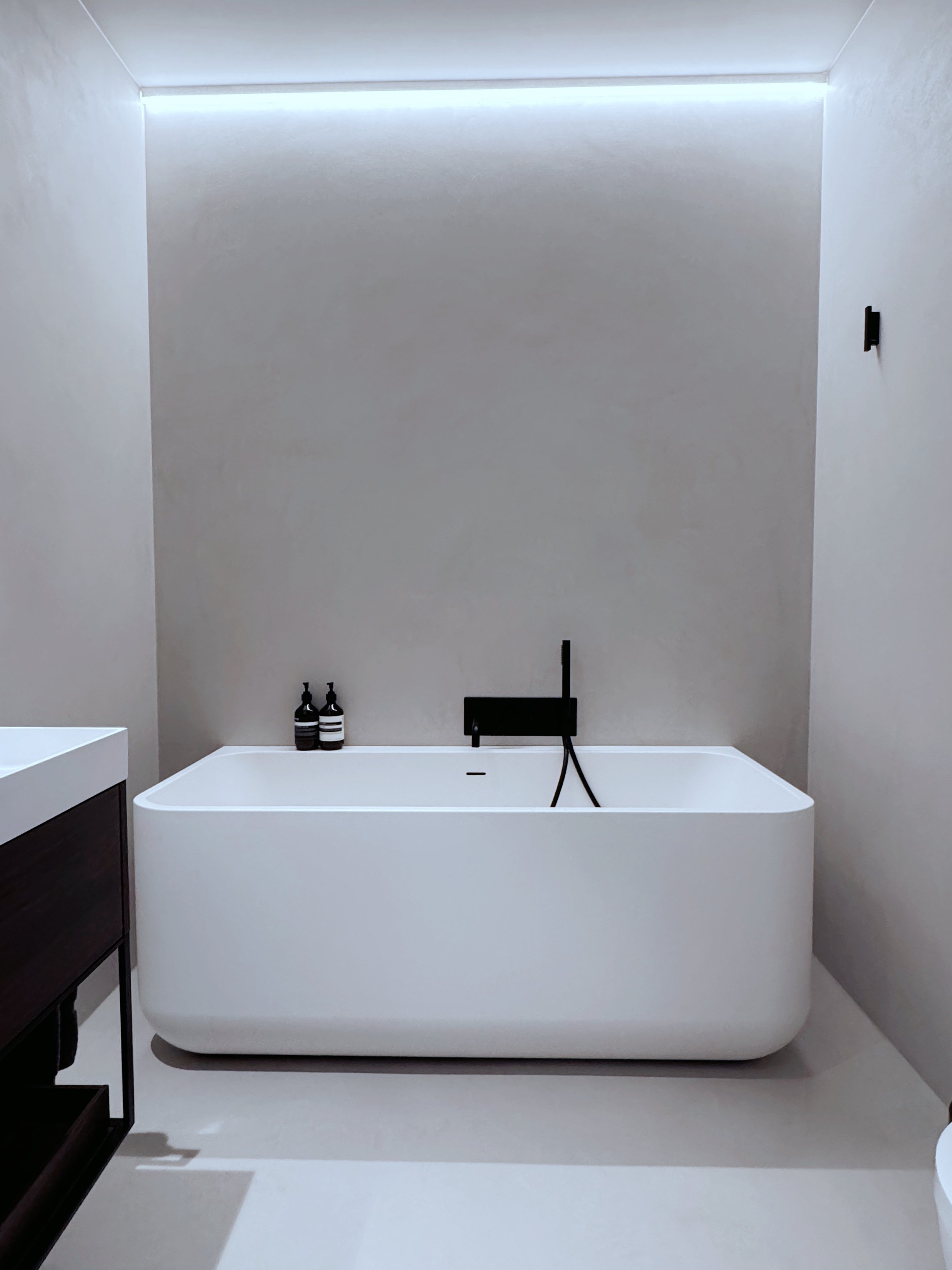 Salle de bain minimaliste avec une baignoire blanche, murs beige clair en micro ciment, et éclairage indirect moderne.