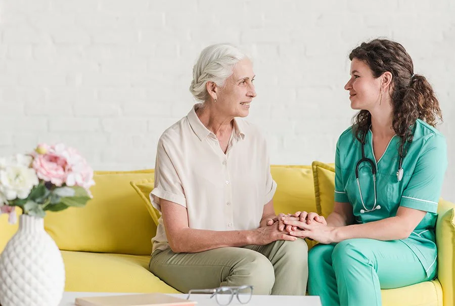 A nurse or healthcare worker wearing teal scrubs and a stethoscope holds the hand of an elderly woman sitting on a yellow couch, both smiling at each other.