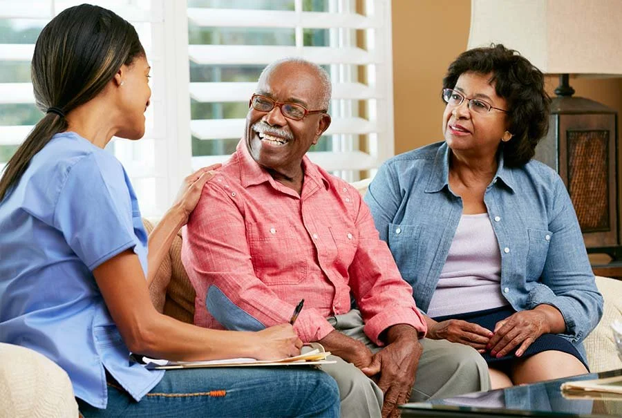 A nurse at a clinic or home health visit talking to an elderly couple, a man and a woman, sitting on a couch, with the nurse smiling and taking notes.