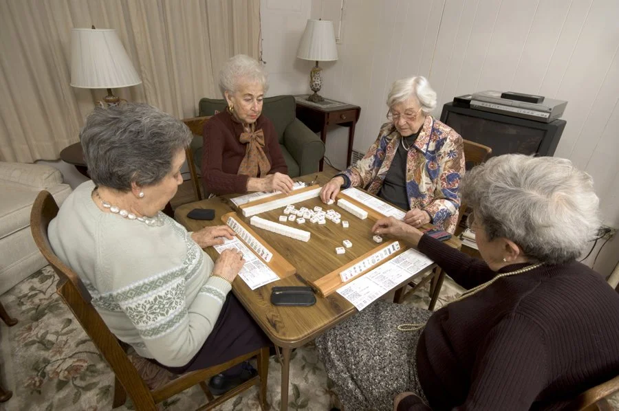 Four elderly women playing a game of Rummikub at a wooden table in a cozy living room.