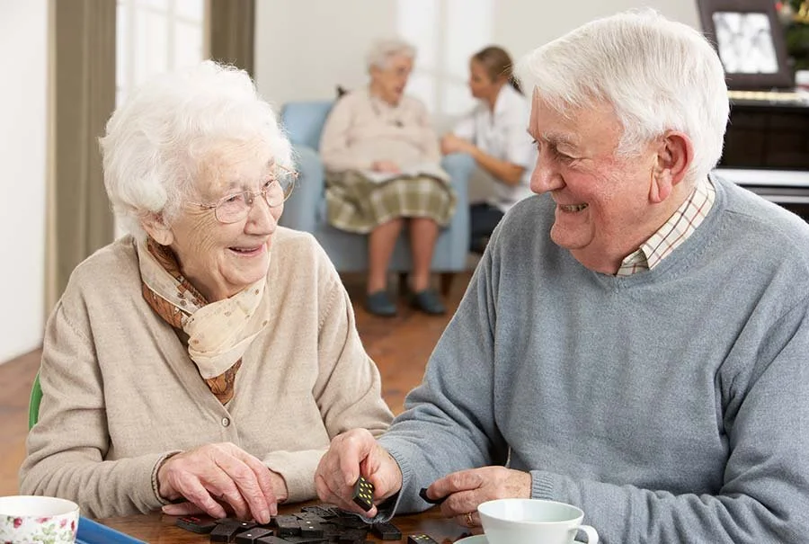 An elderly woman and a man are playing a game with dominoes at a table, smiling and looking at each other. In the background, there is another elderly woman and a caregiver or family member sitting on a sofa, engaging in conversation.