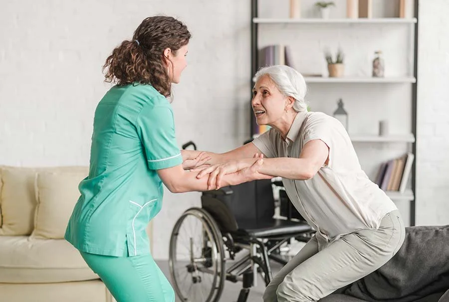 A healthcare worker in a green uniform helping an elderly woman with white hair, sitting on a gray couch, in a bright living room.