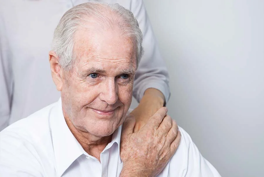 Older man with white hair smiling, receiving a shoulder massage from a person in a white coat, against a plain light-colored wall.