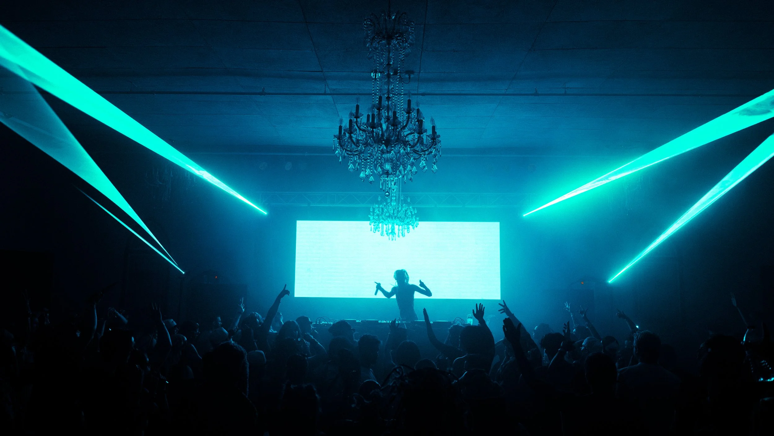 A DJ performs at a concert with blue stage lighting and laser lights, a large LED screen behind them, and an audience dancing with hands raised.