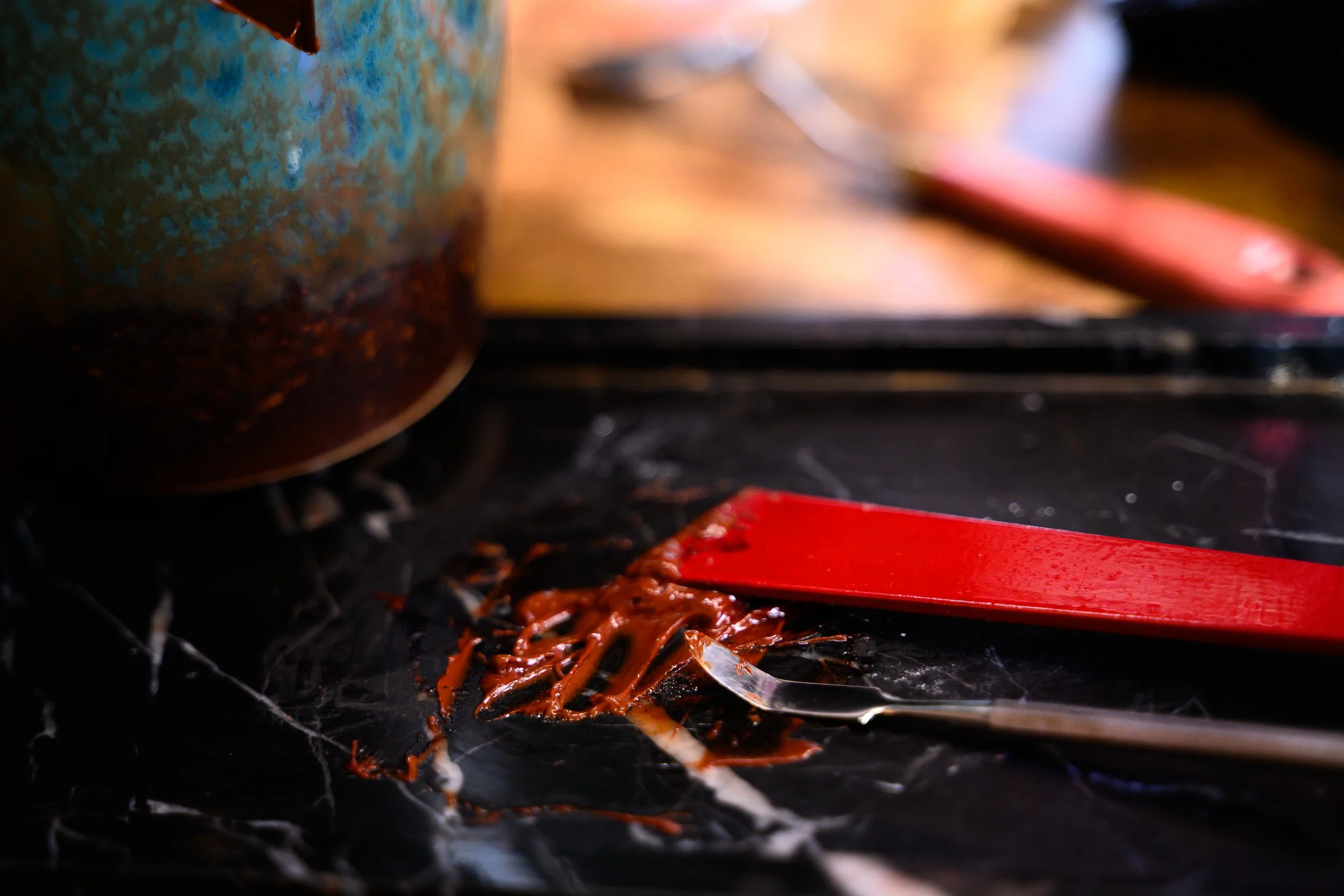A kitchen counter with a spatula and a spoon covered in red sauce, with a pot in the background.