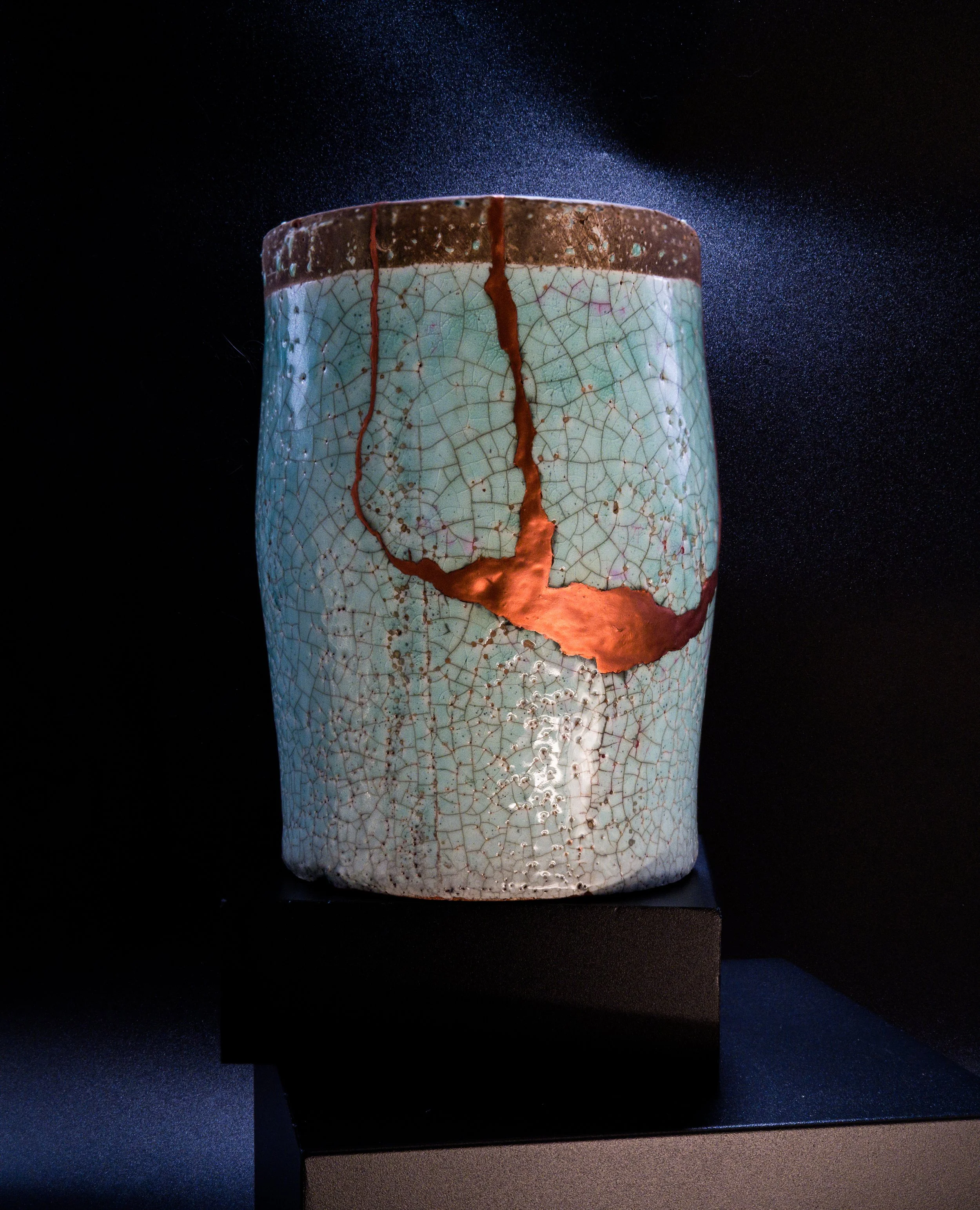 A ceramic vase with crackle glaze and a large broken crack revealing brown clay within, displayed on a black pedestal against a black background.