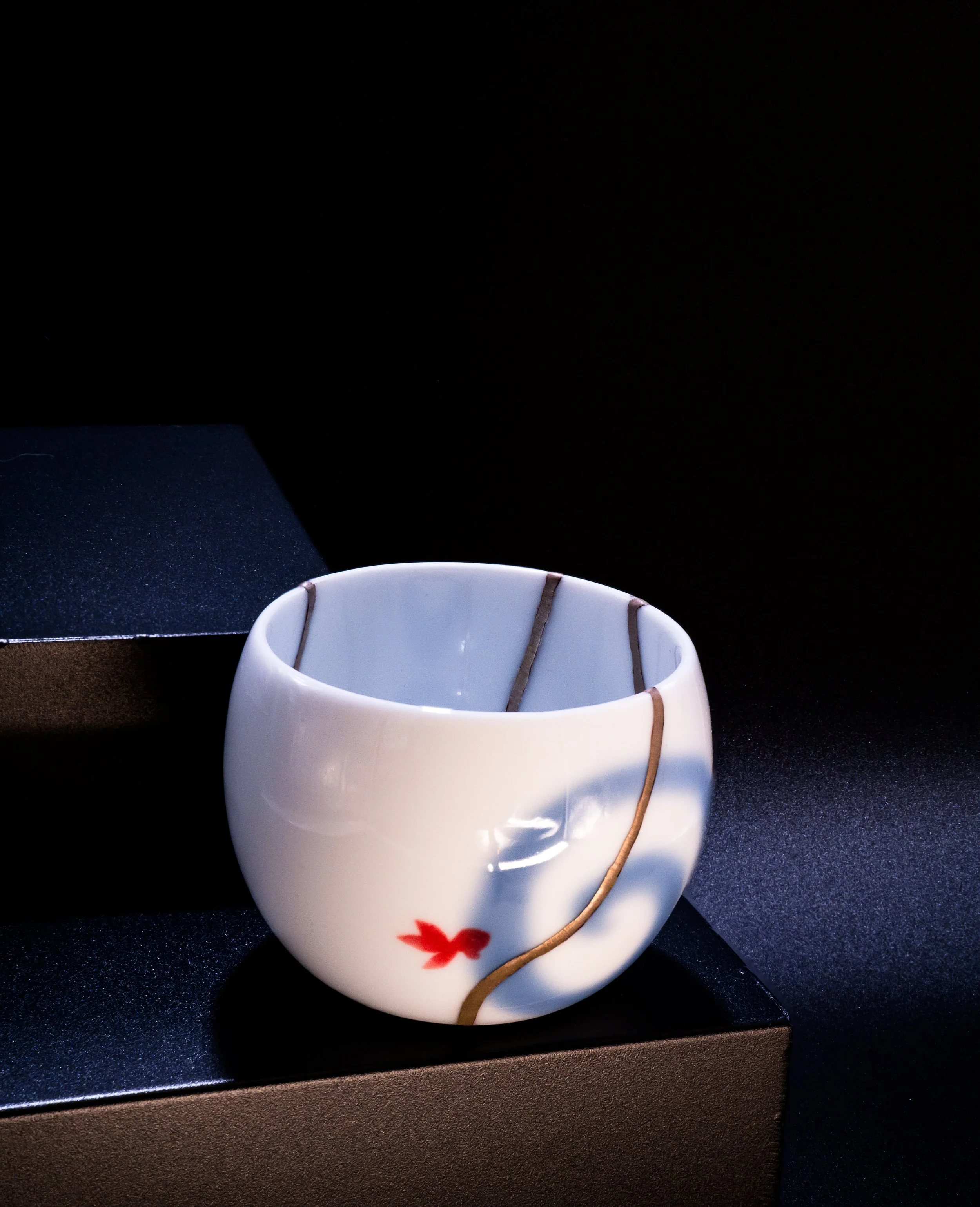 White ceramic cup with a red maple leaf design, decorated with brown and black lines, placed on a black surface with a black background.