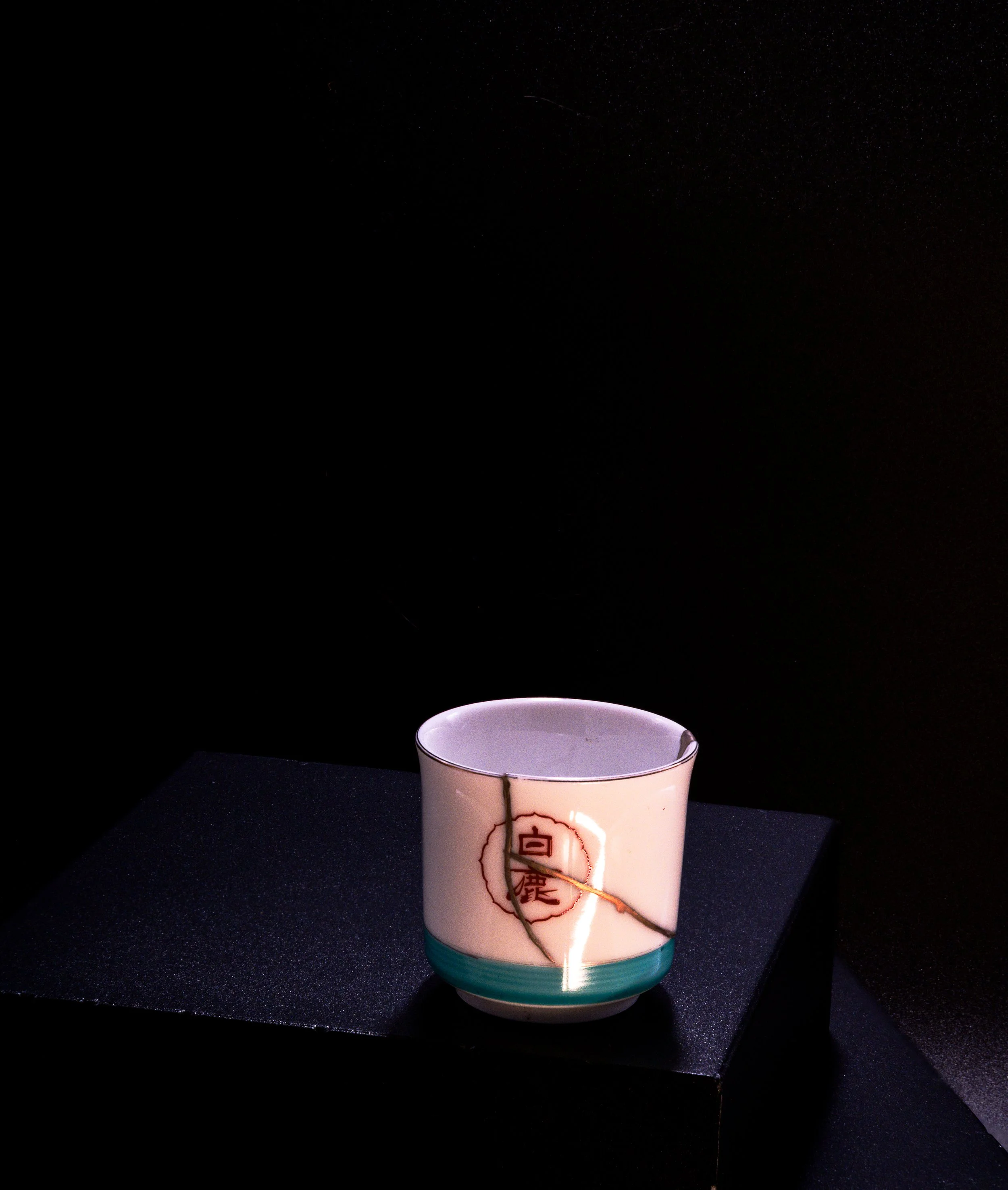 A decorative ceramic cup with a crackle pattern and Japanese characters, displayed on a black pedestal against a dark background.