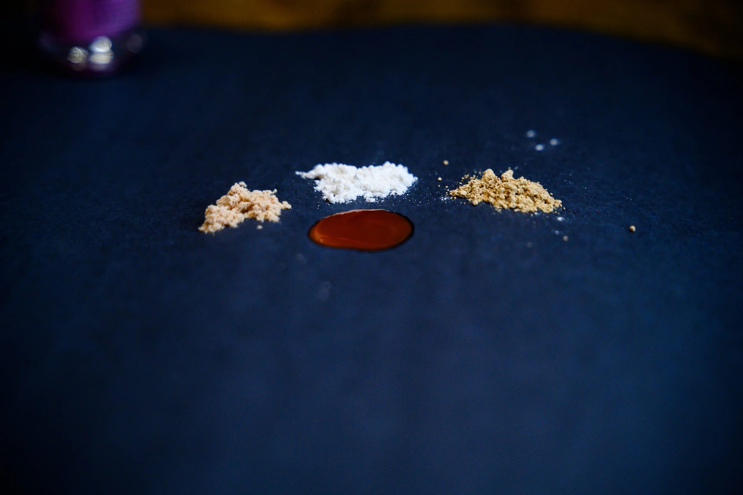 Three small piles of powdery substances—beige, white, and light brown—are arranged in a line on a dark blue surface. A drop of amber-colored liquid is positioned in the middle beneath the powders.