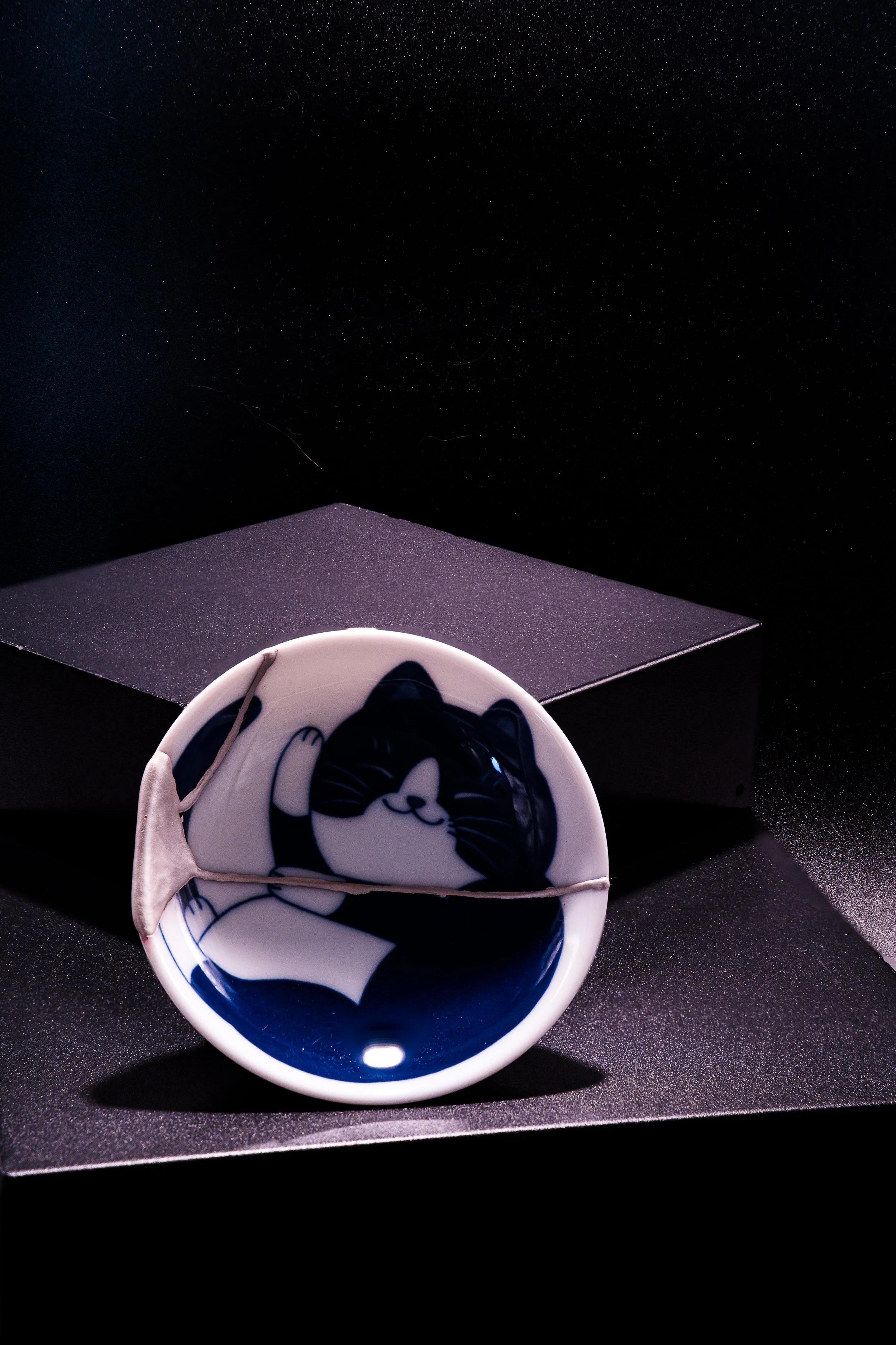 A ceramic bowl with a black and white cartoon cat design, set on a black textured surface with a black box in the background.