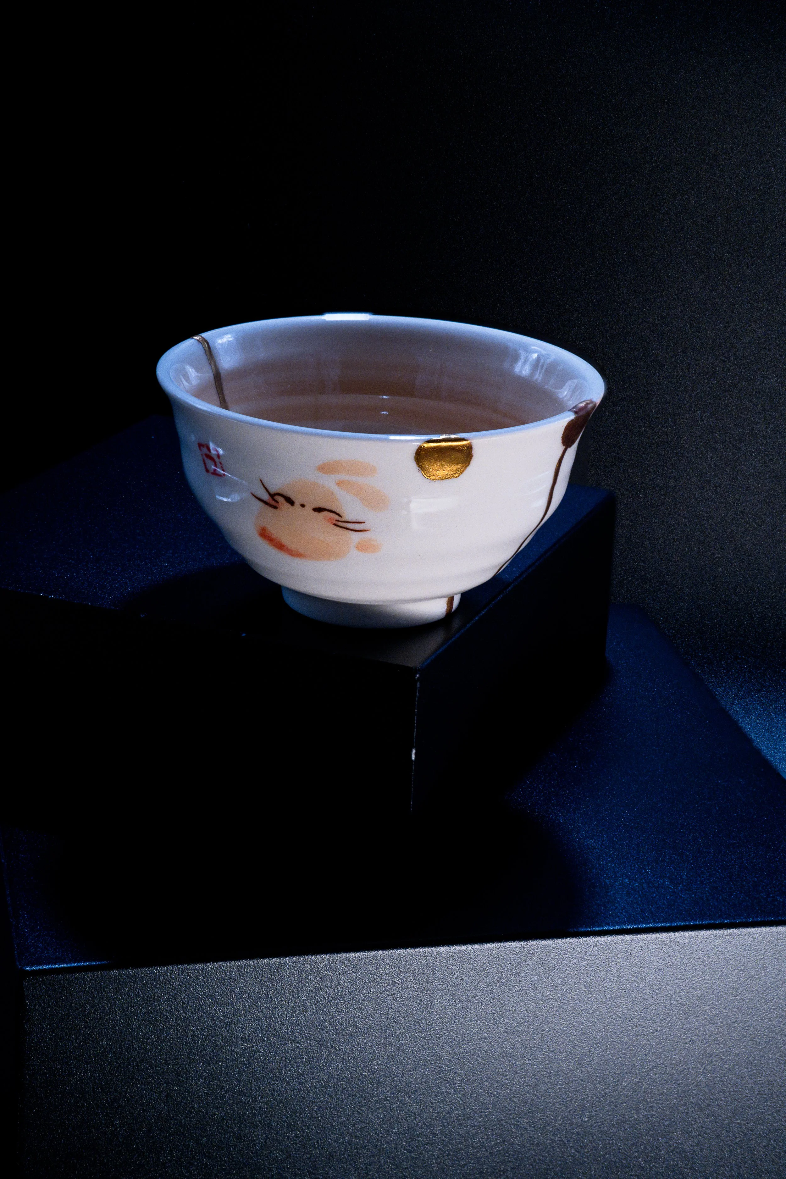 A ceramic bowl, decorated with a face and gold accents, sits on a black pedestal against a dark background.