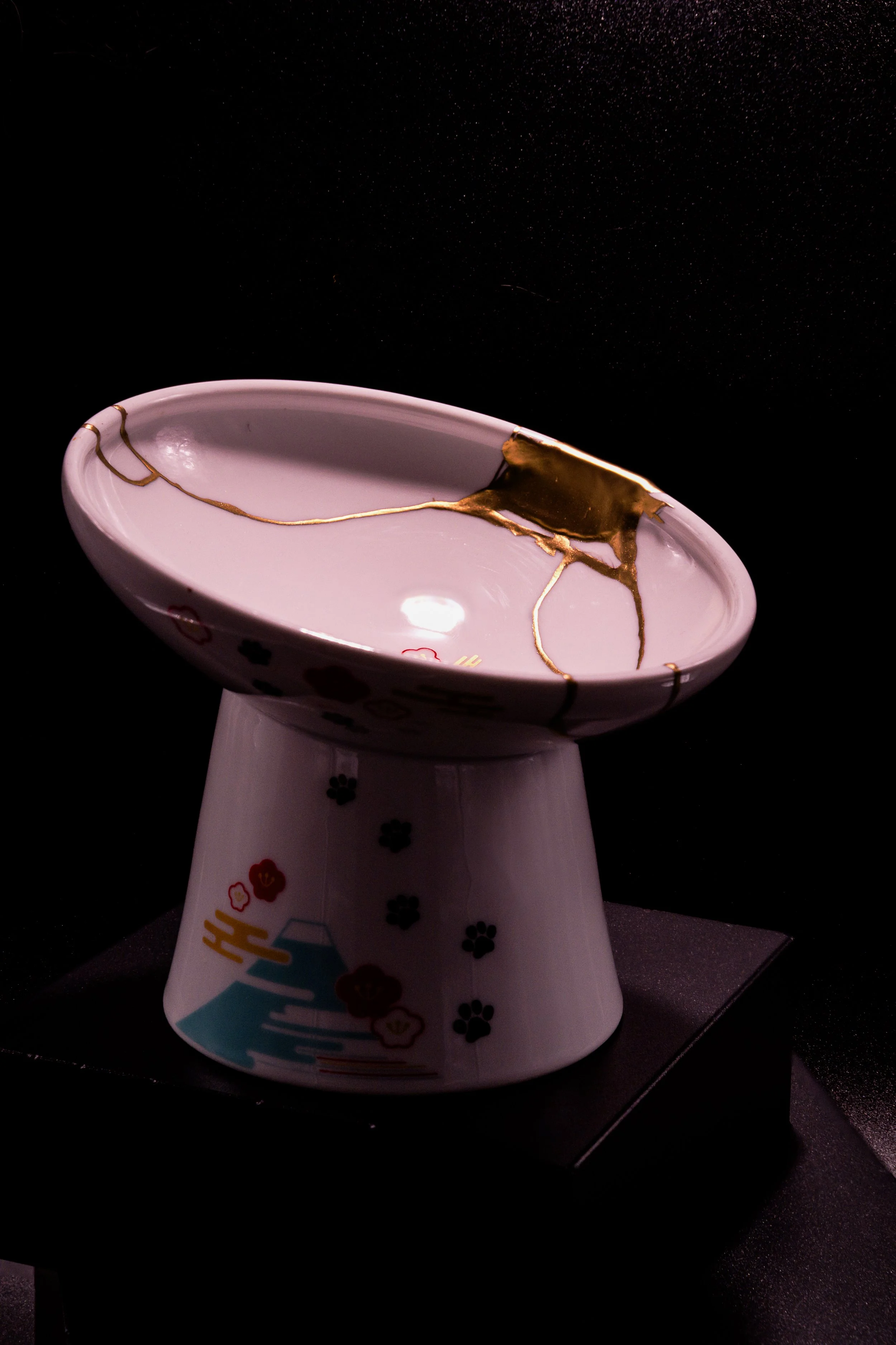 A ceramic bowl with a traditional Japanese kintsugi repair, featuring gold cracks, mounted on a matching pedestal with decorative motifs, displayed on a black surface against a dark background.