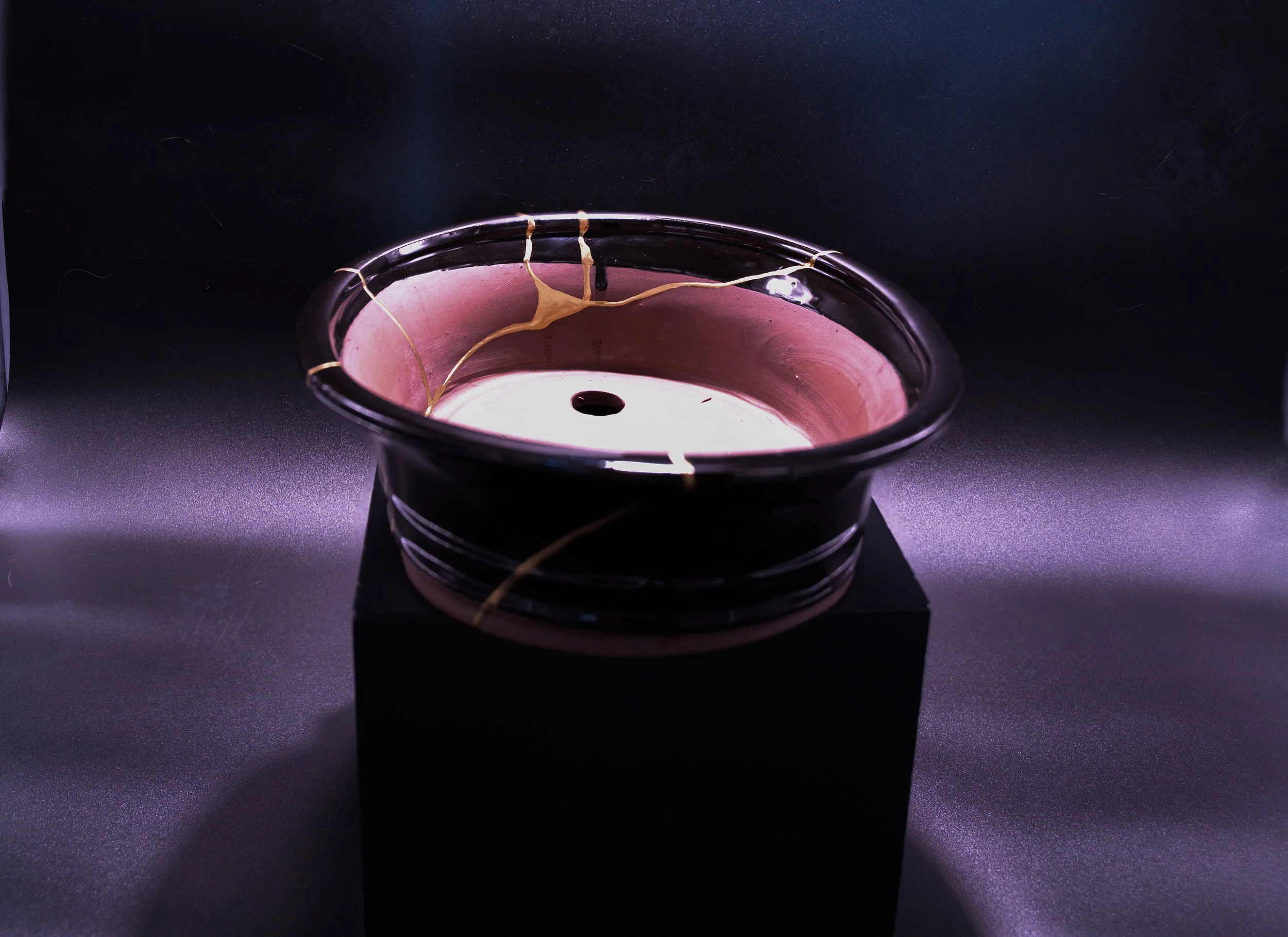 A ceramic or resin sculpture with cracks, pink and white interior, and black exterior, placed on a black pedestal against a dark background.