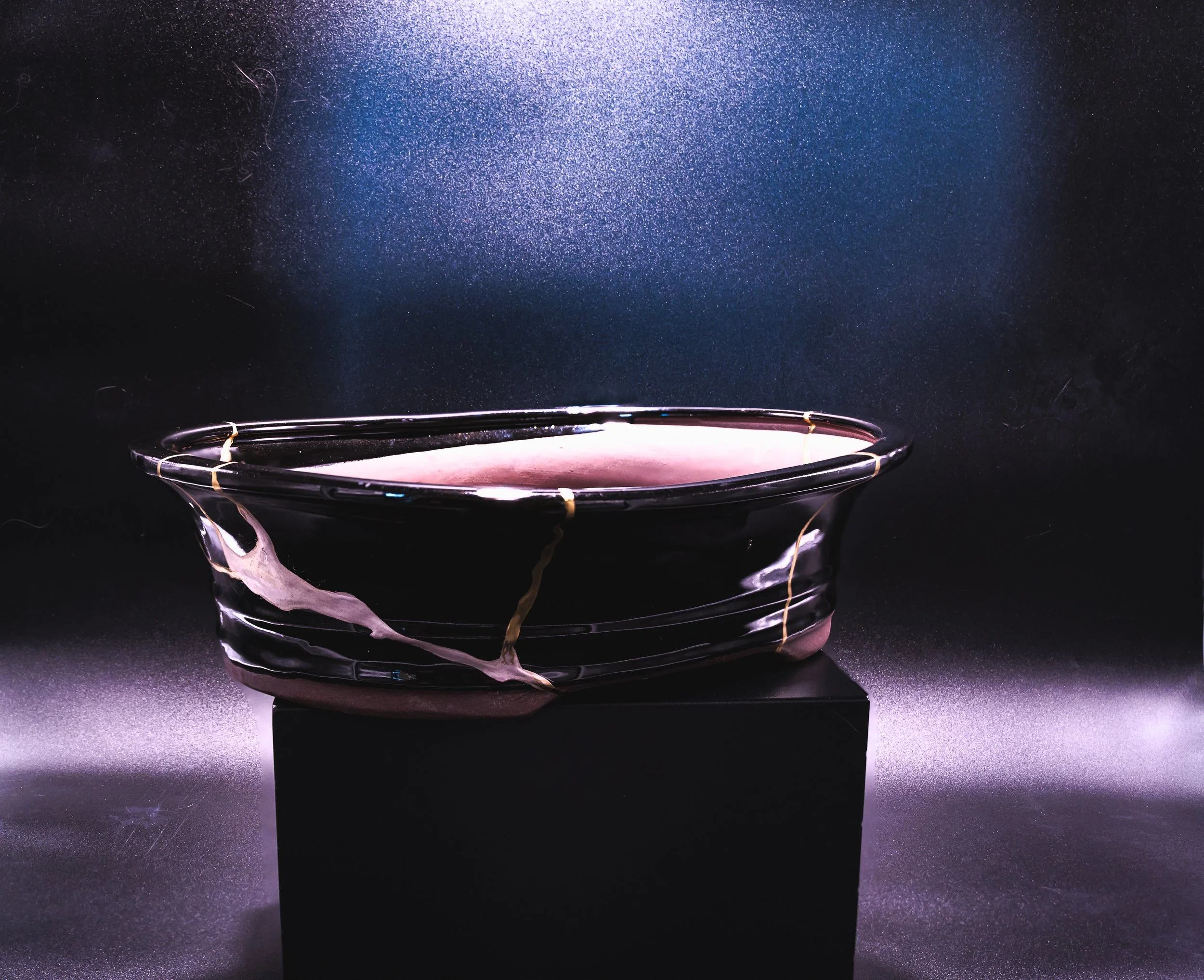 A translucent, black and pink glass bowl with decorative yellow bands, sitting on a black pedestal against a dark, textured background with blue accents.