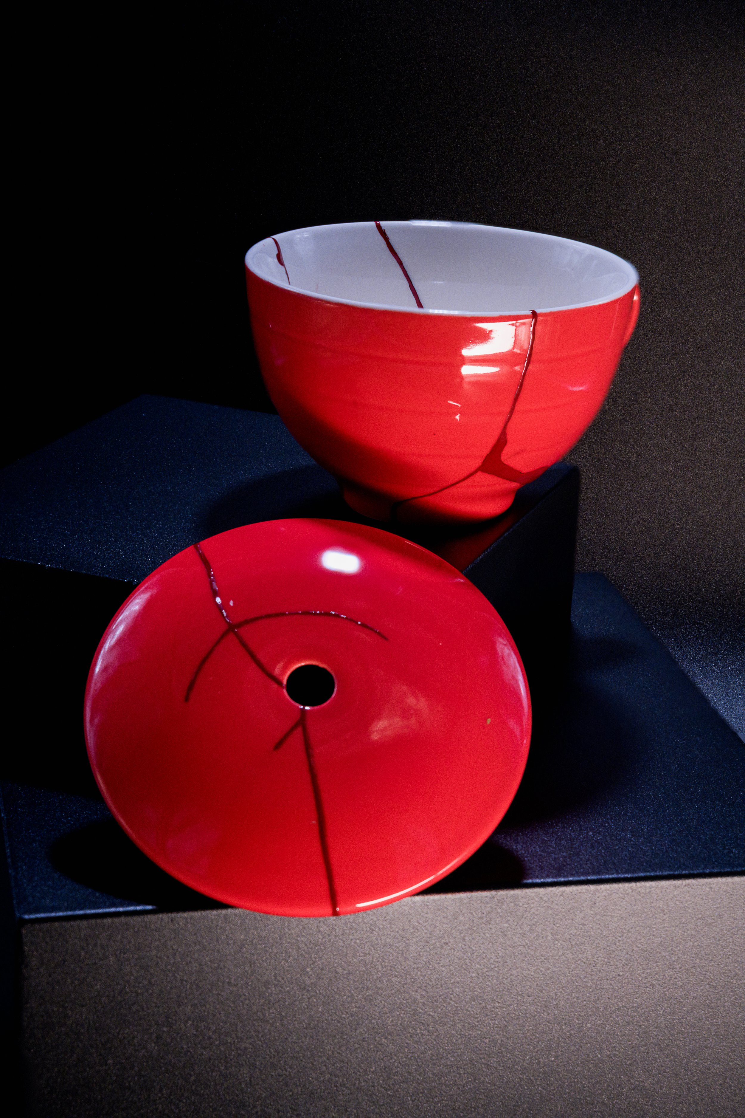 A red and white ceramic bowl with black lines resembling a crack pattern, displayed on a black pedestal against a dark background.