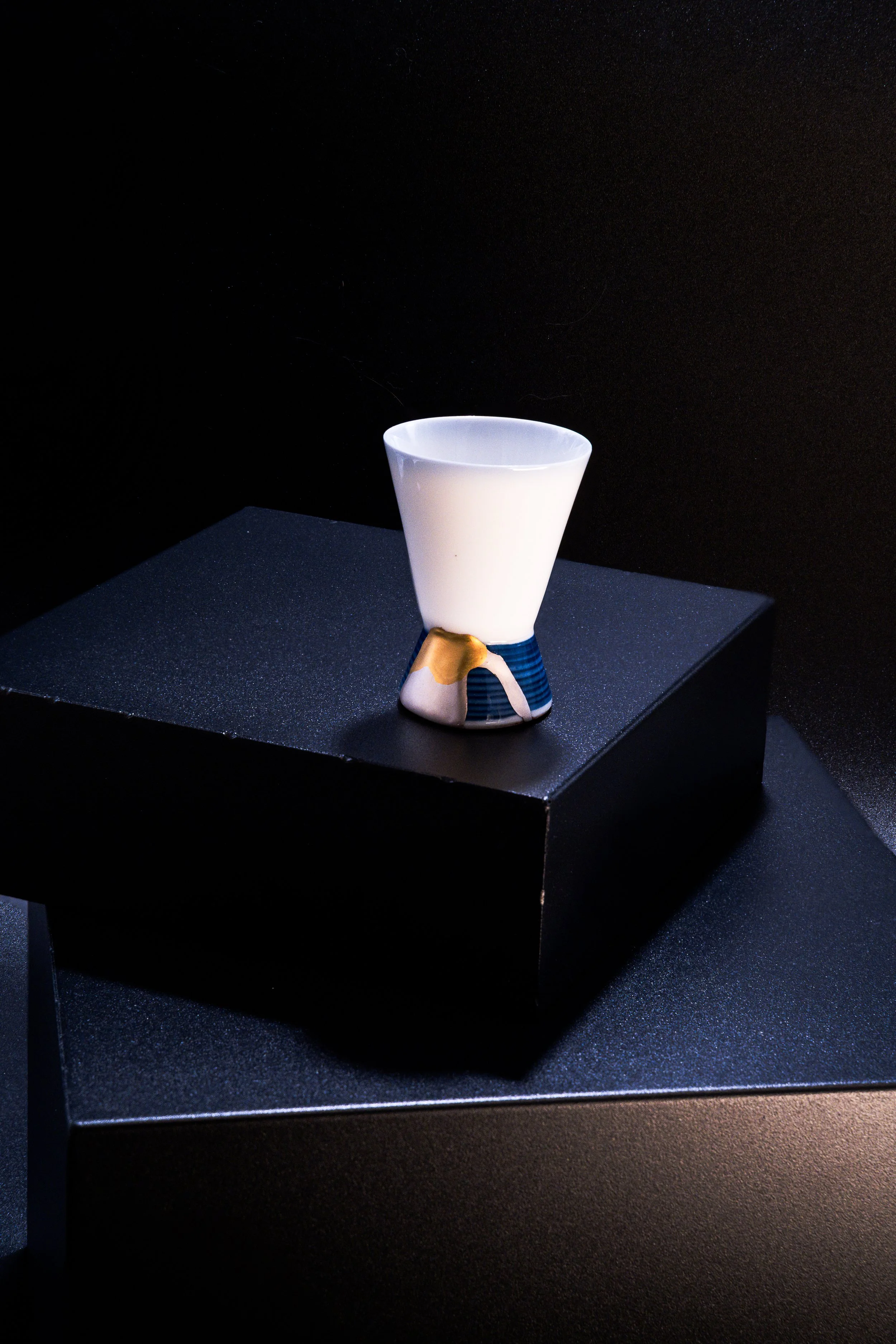A white ceramic cup with a unique handle design, displayed on a black platform against a dark background.