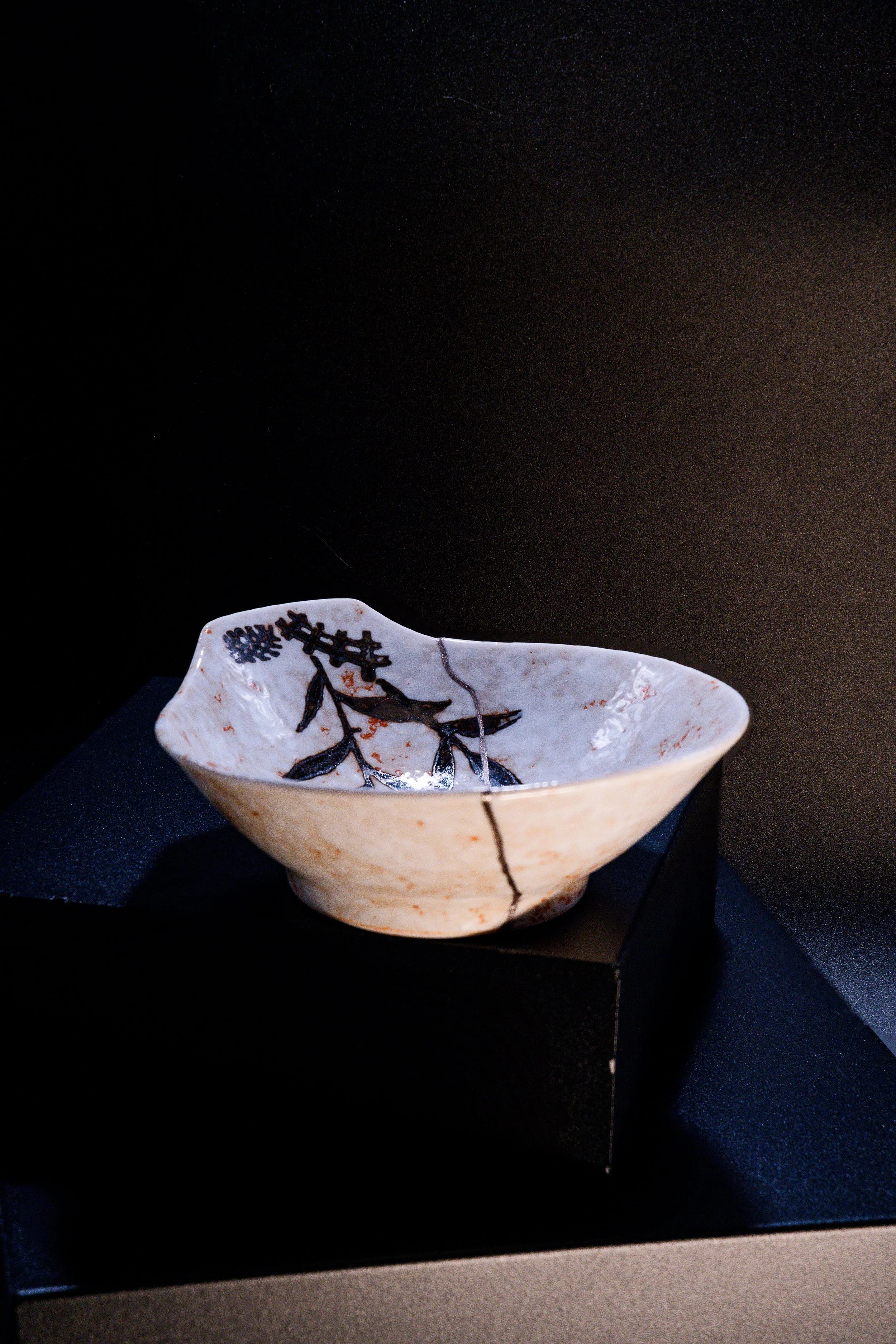 A small, decorative ceramic bowl with a black floral design on a white background, displayed on a black stand against a dark backdrop.