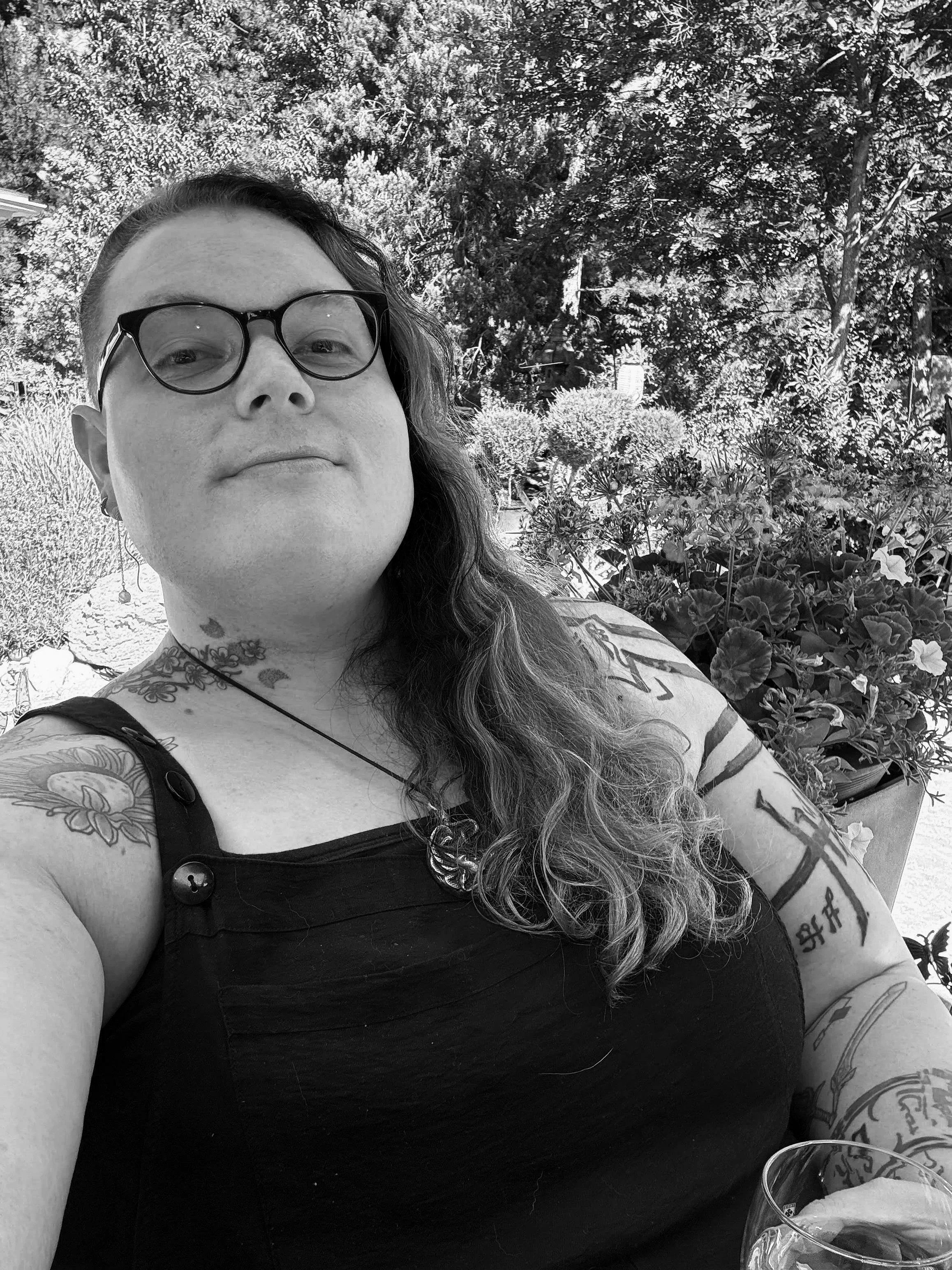 A woman with long, wavy hair, glasses, and tattoos on her arms and shoulder, sitting outdoors with plants and trees in the background.