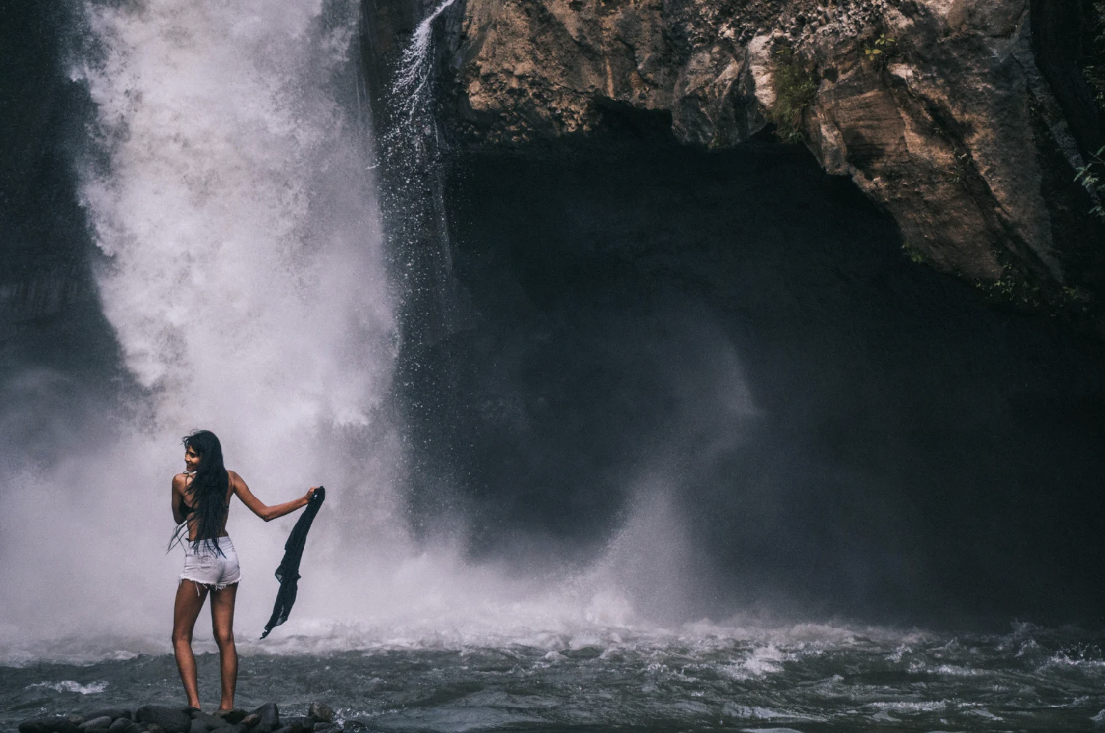A woman standing on rocks near a waterfall, holding a piece of clothing, with water splashing behind her.