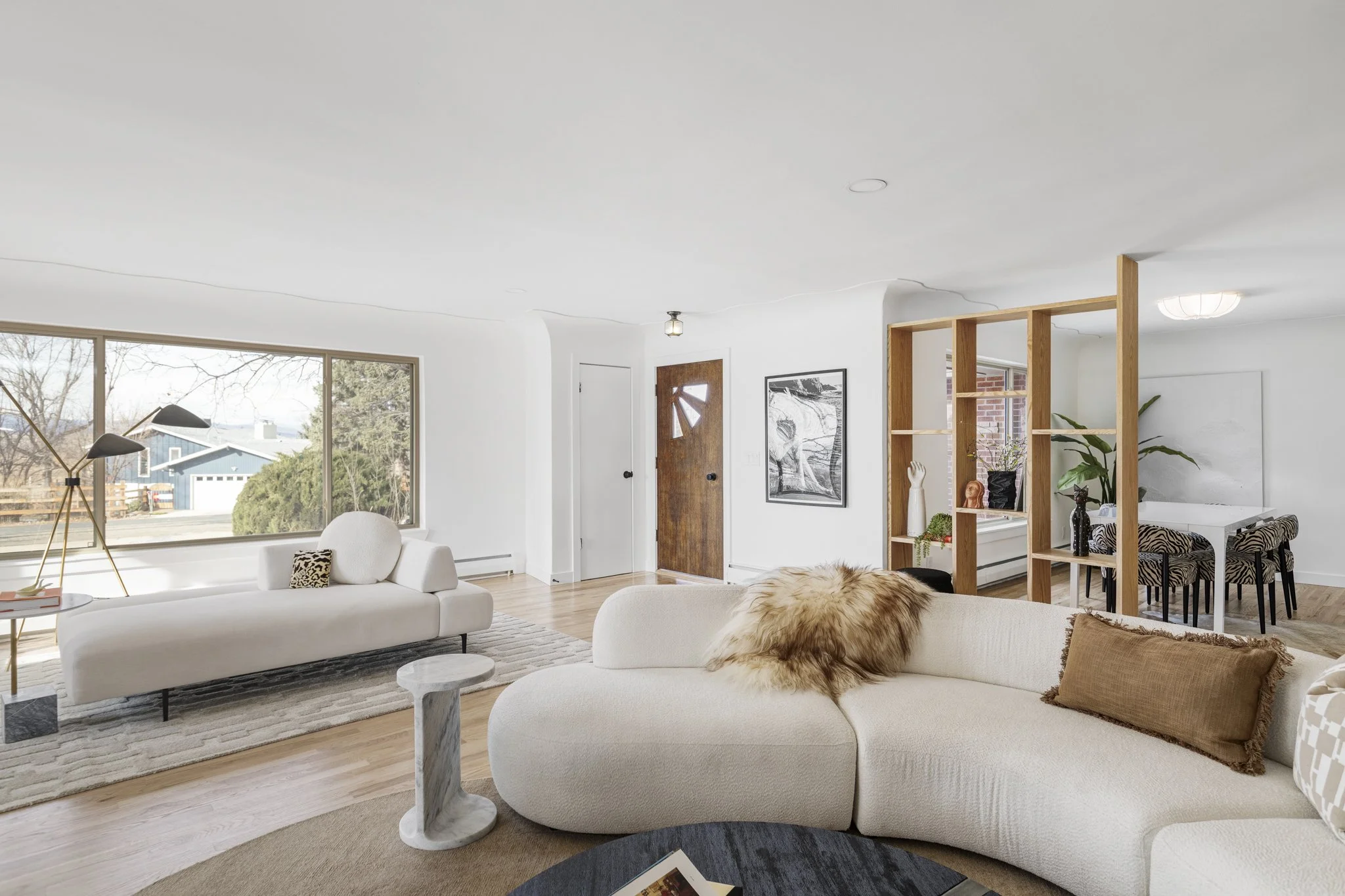 Mid-mod bright living room with large window, white sofa, modern floor lamp, wooden room divider, dining area with zebra print chairs, and decorative plants.