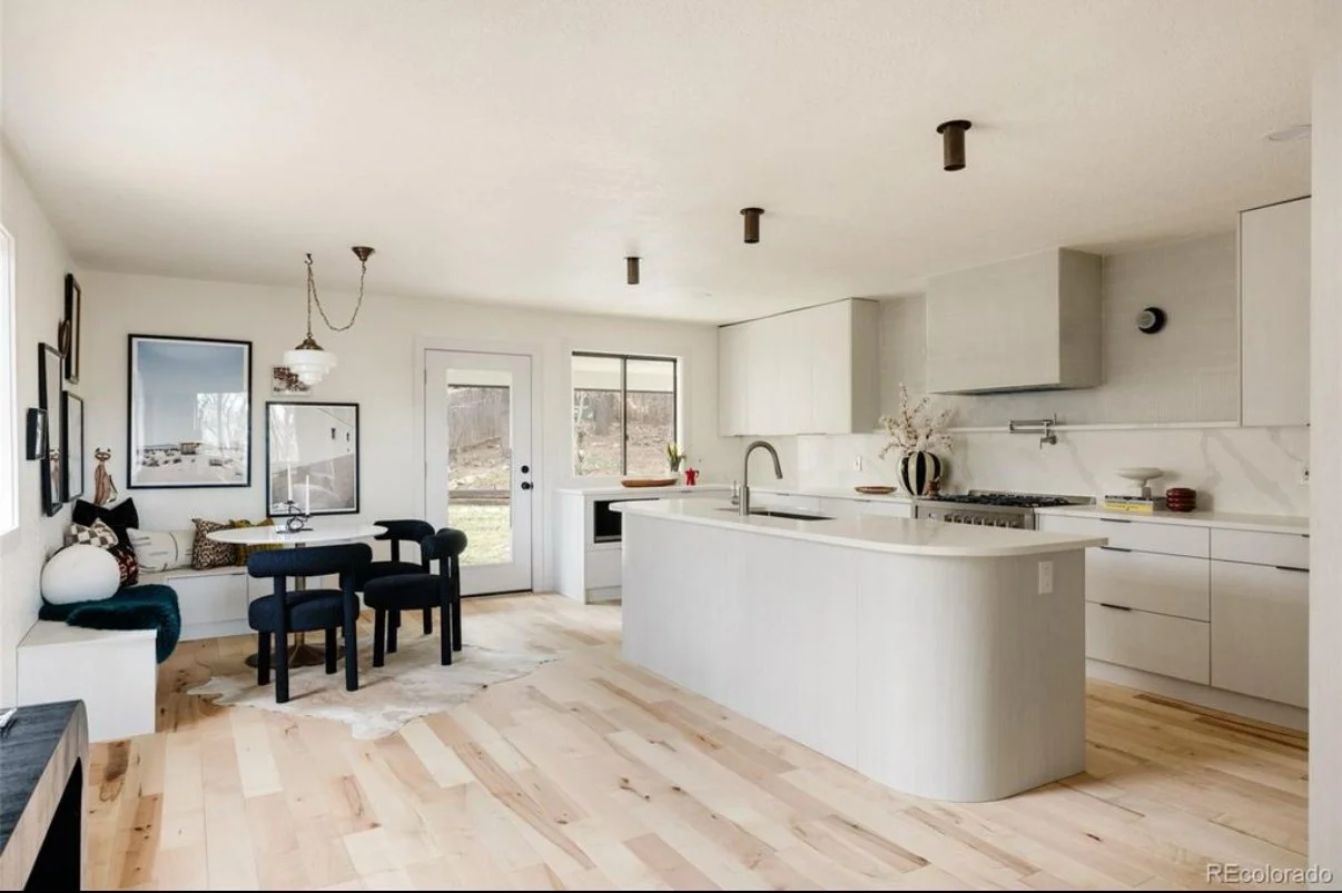 Bright, minimalist kitchen and dining area with white cabinets, countertops, walls, a small dining table with three black chairs, a window, a door, decorative wall art, and light wood flooring.