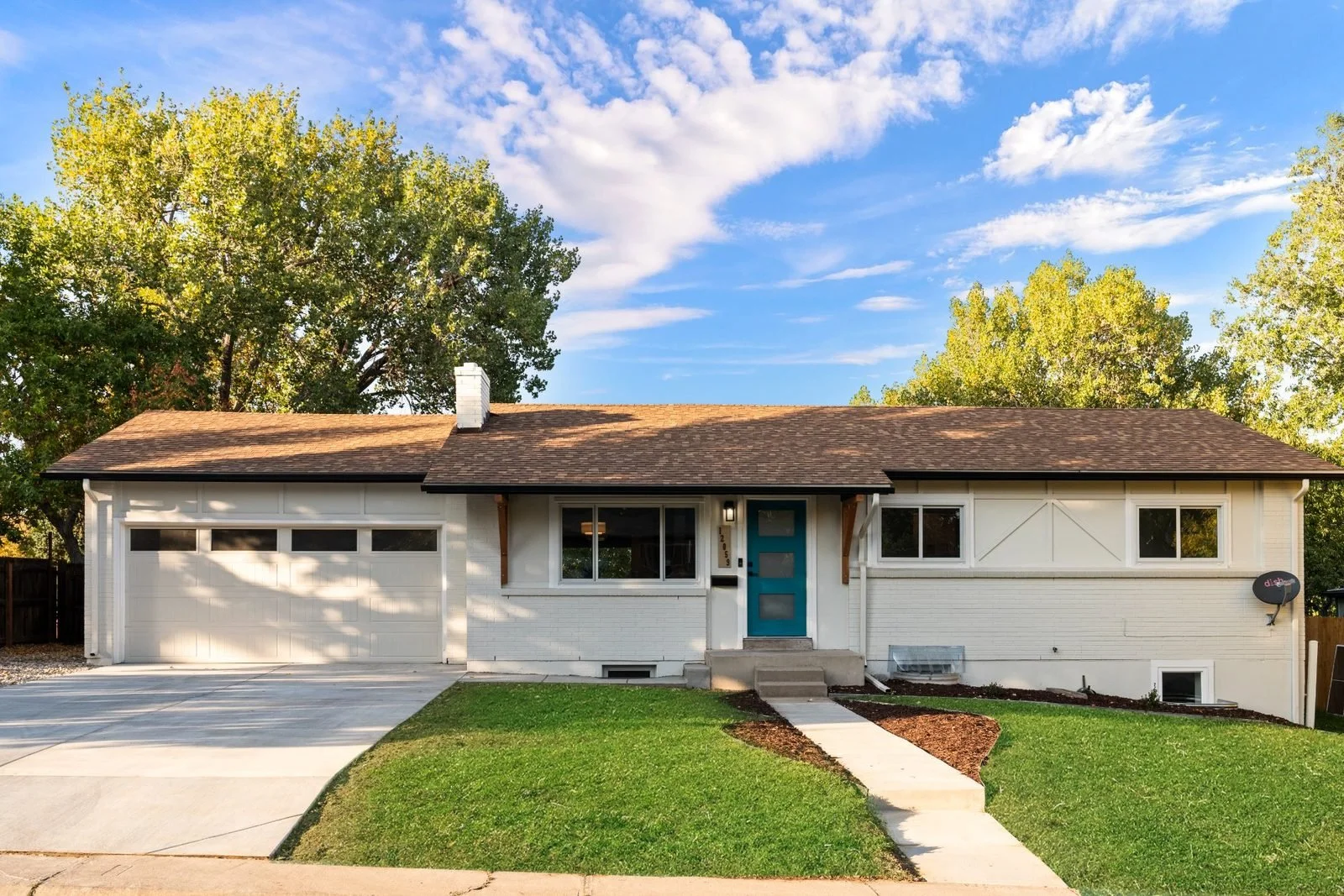 A single-story house with a white brick exterior, a blue front door, two car garage, and a front yard with a concrete walkway, green grass, and large trees in the background under a partly cloudy sky.