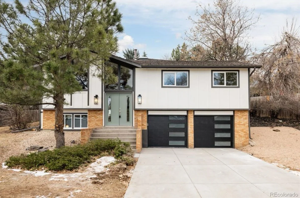 Modern two-story house with a brick and white exterior, black garage doors, and front steps leading to a green front door. There are large windows and trees, with a driveway and a small landscaped area in the front.