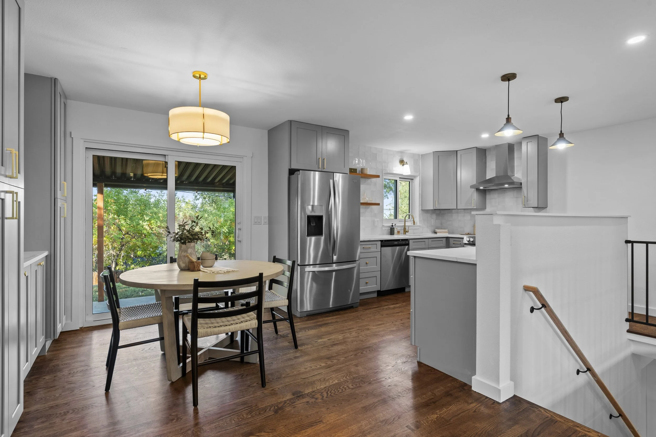 Modern kitchen with gray cabinets, stainless steel appliances, a round dining table with four chairs, wood flooring, and a view of green trees through sliding glass doors.
