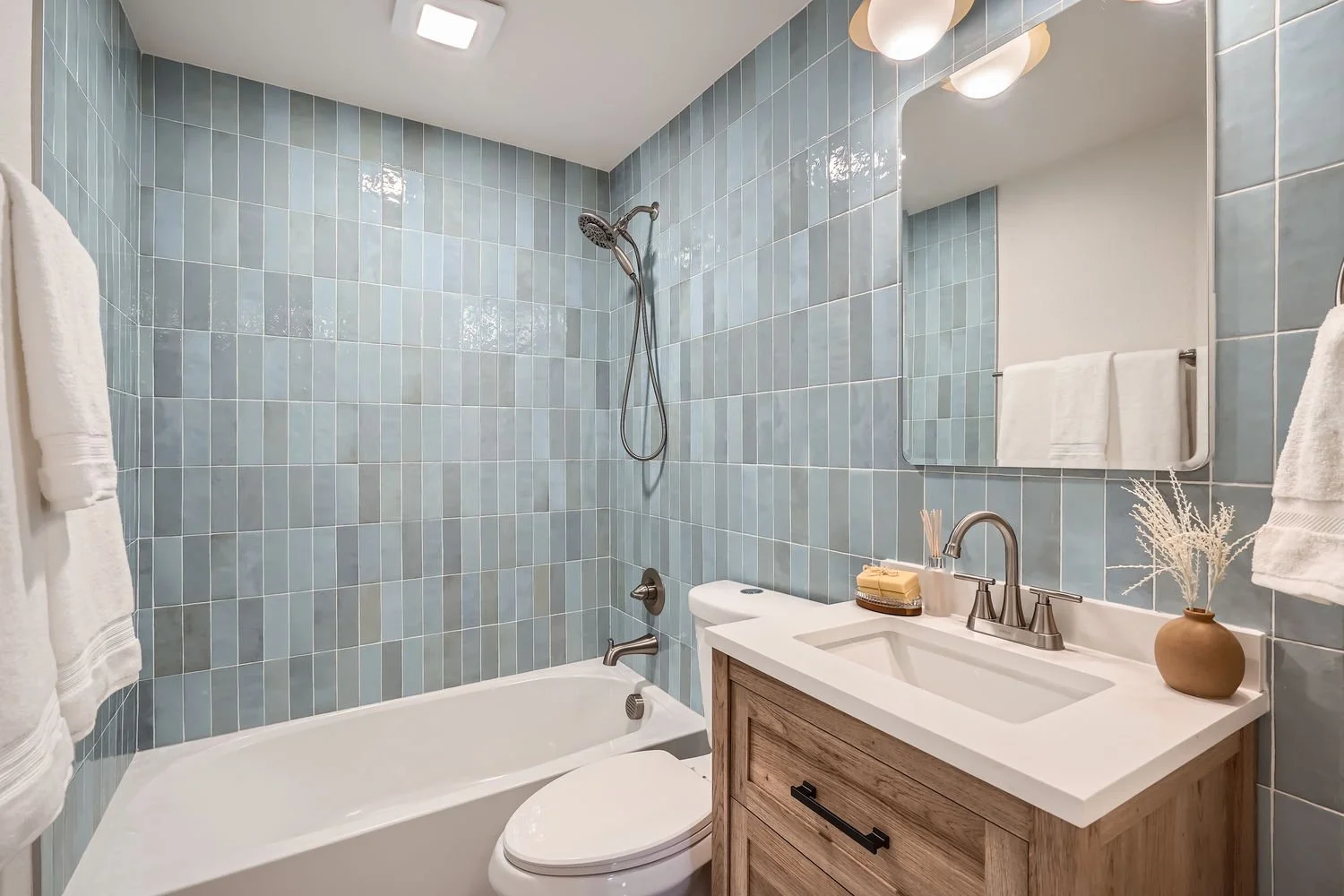 Bathroom with blue tiled wall, a bathtub with a shower head, a toilet, a wooden vanity with a white sink, a mirror, and decorative items including a vase with dried plants and towels.