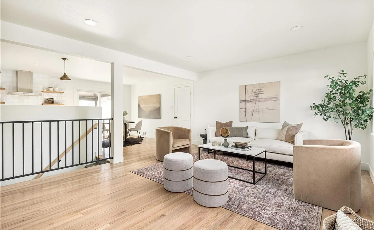 Living room with white walls, light wood flooring, and modern furniture including a white sofa, beige chairs, and a black coffee table. Decorative items such as plants, art, and a rug are present.