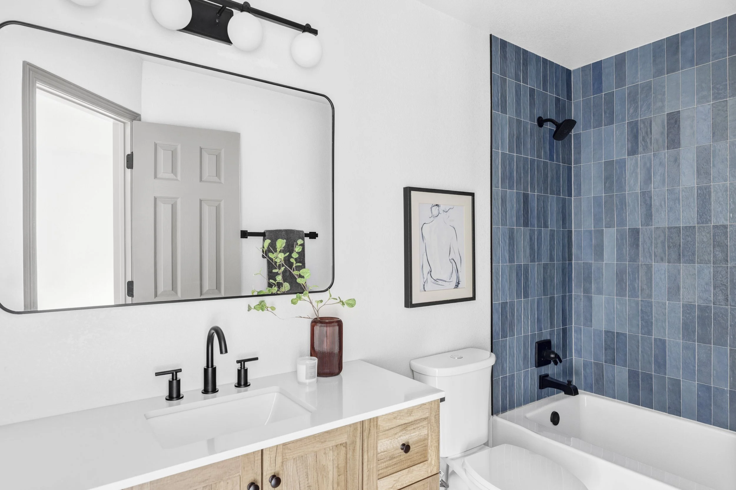 Bathroom with white walls, a mirror above a white sink with black fixtures, a wooden vanity, a vase with a green plant, a framed line drawing, a white toilet, and a shower area with blue tiles and black fixtures.