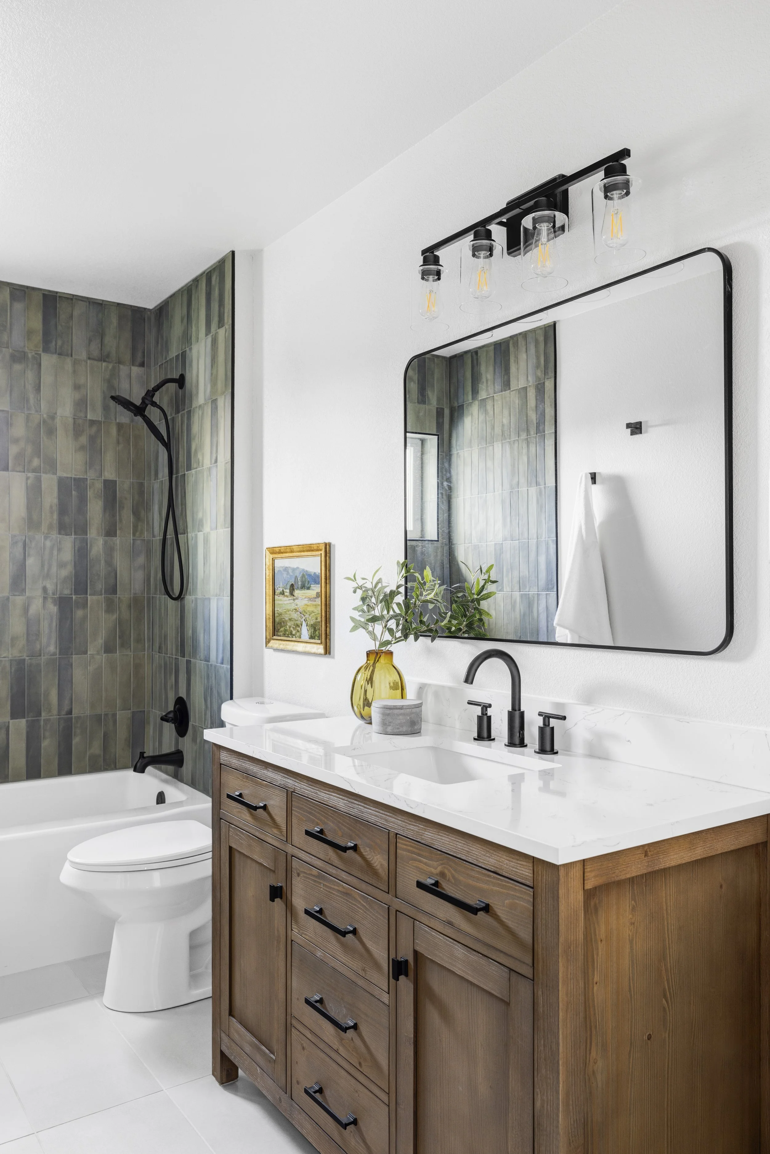 Modern bathroom with wooden vanity, white countertop, and black fixtures. Includes a mirror, a plant in a yellow vase, and a framed landscape painting. Shower area with dark tiles and a black showerhead.