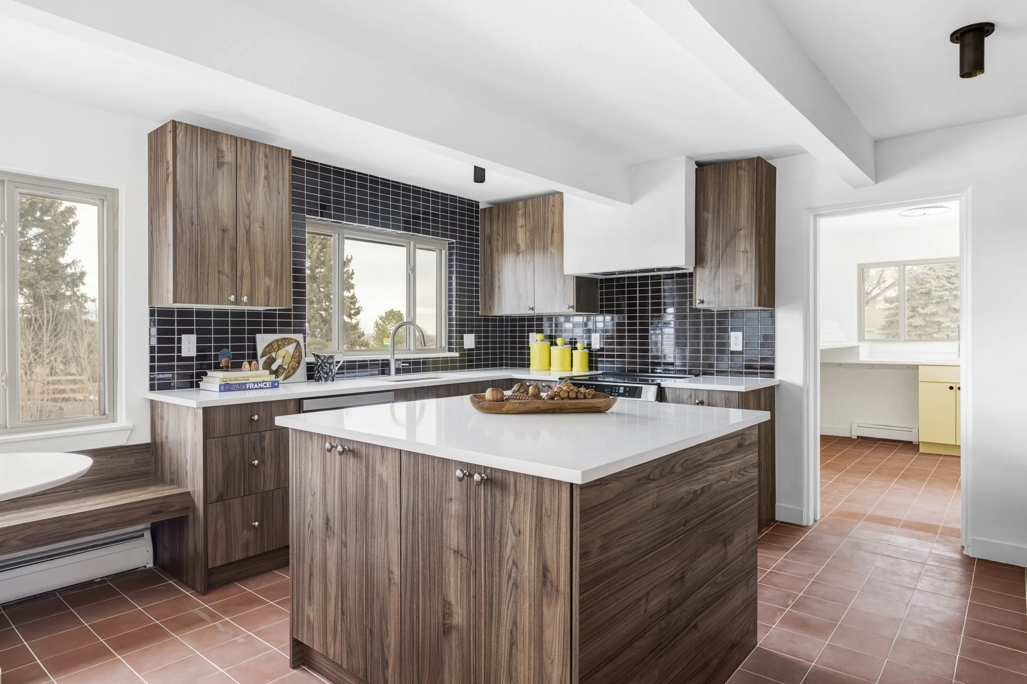 Mid-Mod kitchen with wooden cabinets, black tiled backsplash, white countertops, and terracotta floor tiles, with a window over the sink and a doorway leading to a bright but darker adjacent room.