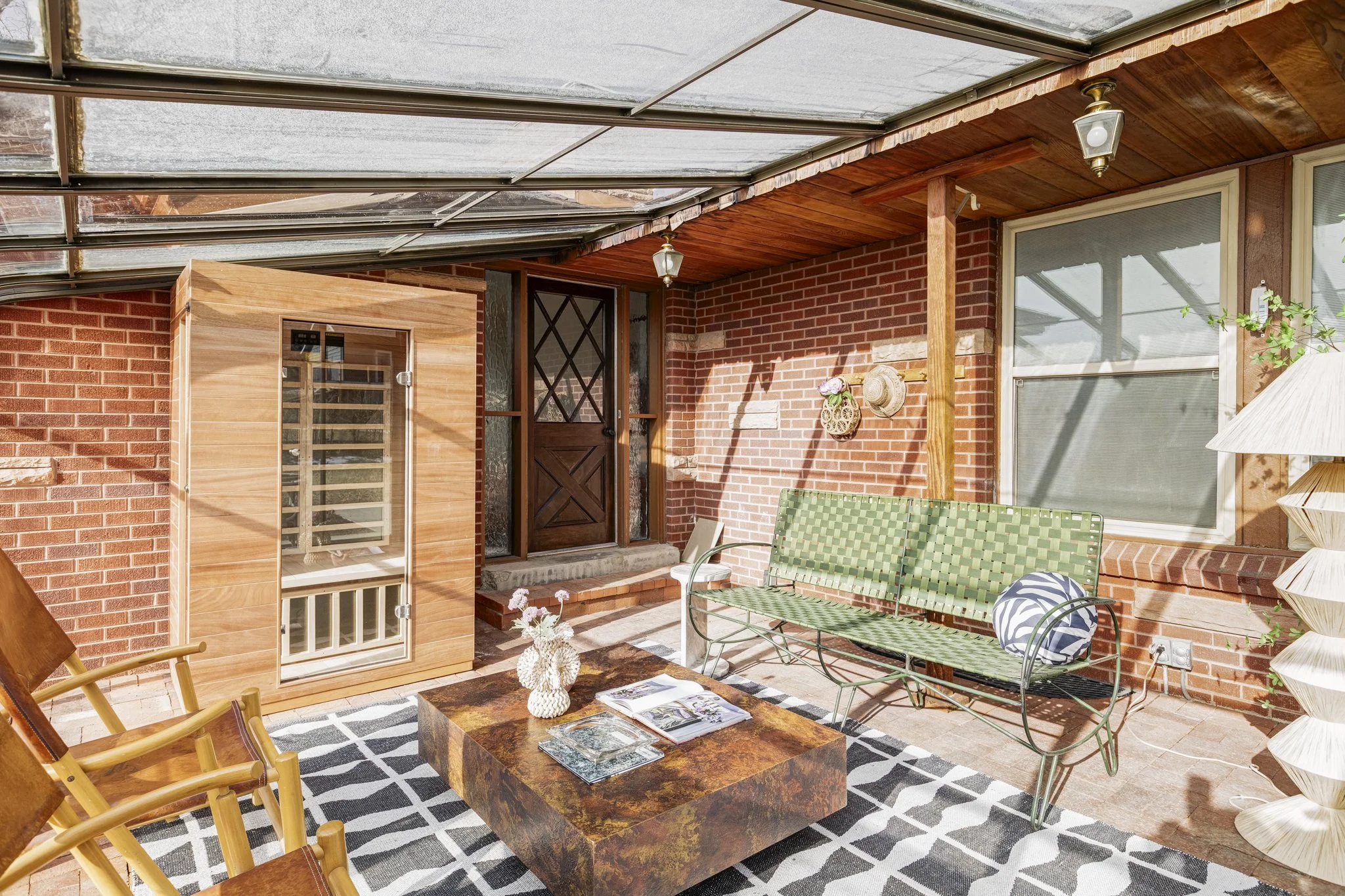 Sunlit enclosed porch with brick walls, wooden ceiling, and large windows. Features a green metal bench with a round pillow, a wooden rocking chair, a marble-topped table with books and a decorative vase, and a wooden sauna cabinet.