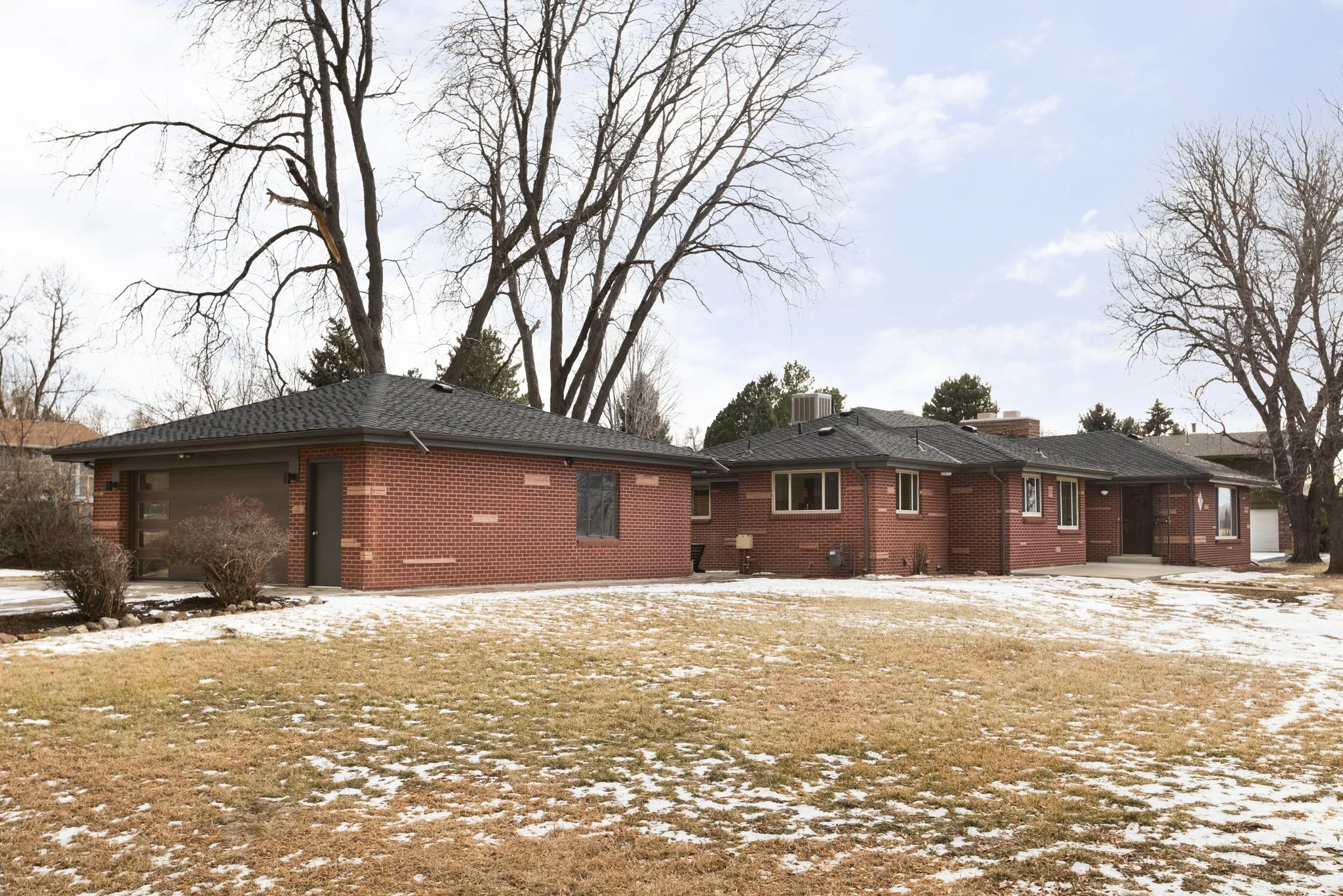 Mid-mod brick house with a black roof, several windows, and a small porch, situated in a yard with brown grass, leafless trees, and patches of snow under a cloudy sky.