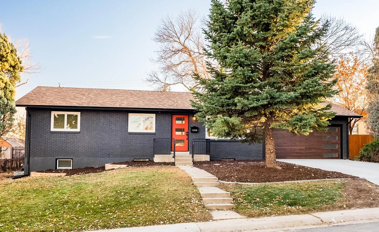 A modern single-family house painted dark gray with a red front door, a large evergreen tree in the front yard, and a concrete driveway leading to a garage.